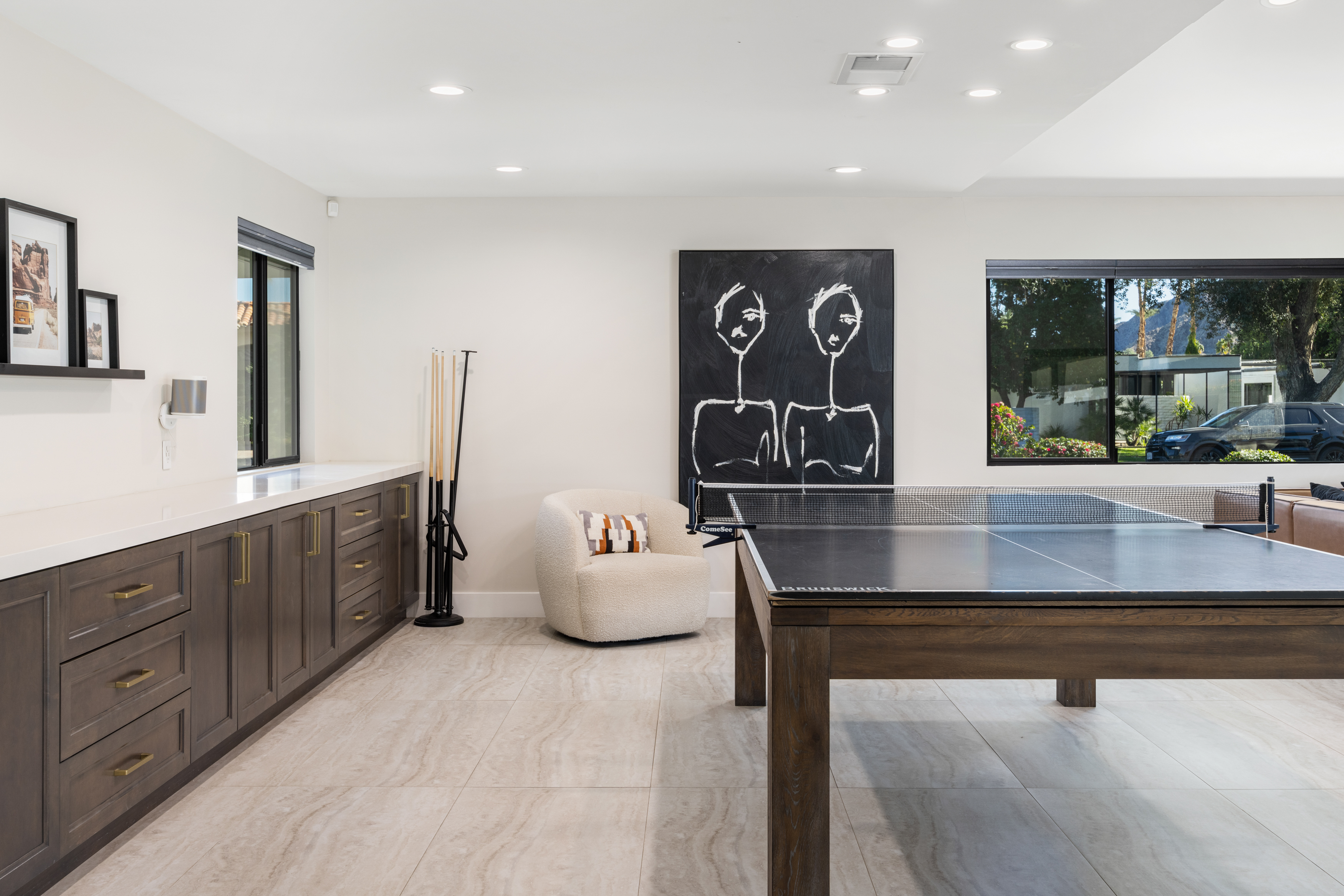 This bright basement features light tile flooring, built-in cabinetry with a white countertop, and an abstract art piece. A ping pong table dominates the space, suggesting a recreational area. The room's design is simple yet stylish, offering a versatile space for entertainment or relaxation.