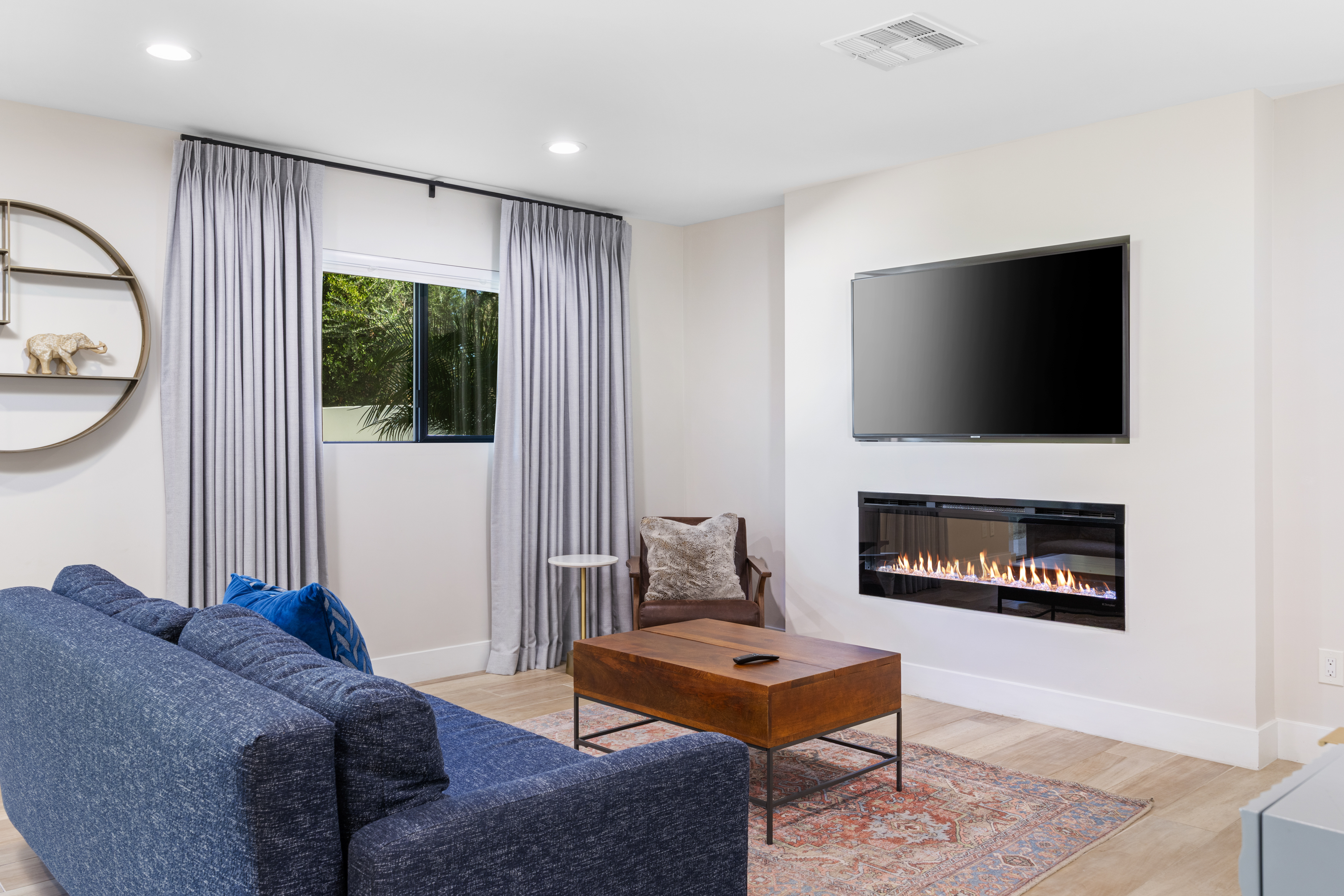 This is a well-lit living room featuring a modern fireplace and TV mounted above it. A comfortable blue sofa faces a wooden coffee table on a patterned rug. The room also includes a window with curtains and a decorative shelf, offering a welcoming and stylish space.