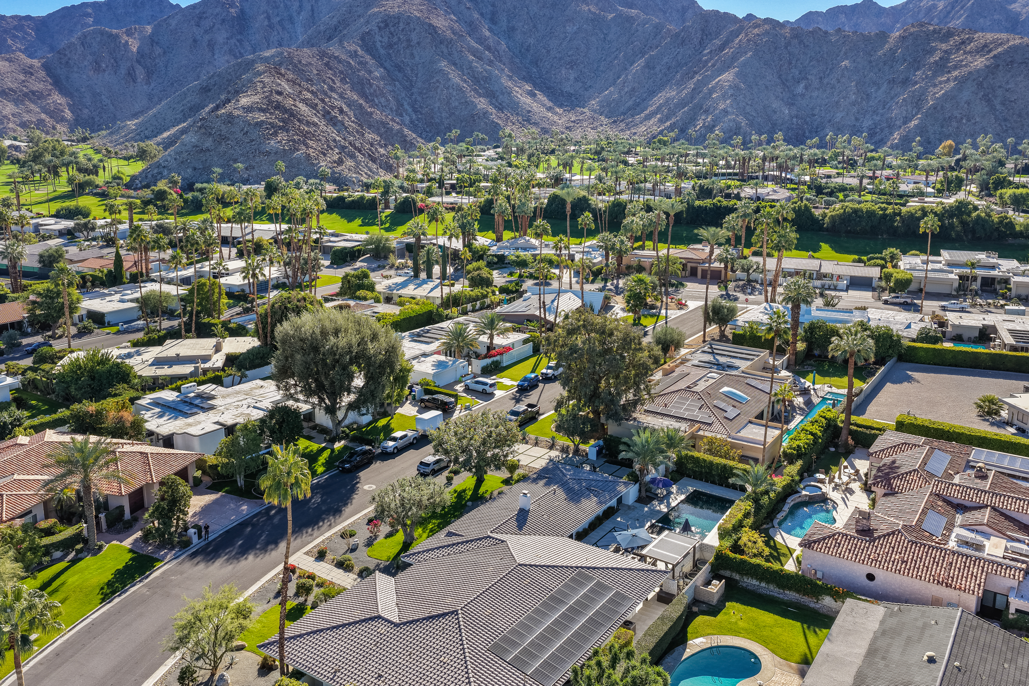 This aerial view captures a luxurious residential neighborhood nestled against a mountainous backdrop. The scene features well-manicured lawns, mature trees, and private swimming pools, creating an impression of upscale suburban living. Solar panels are visible on some rooftops, denoting an environmentally conscious approach to homeownership.