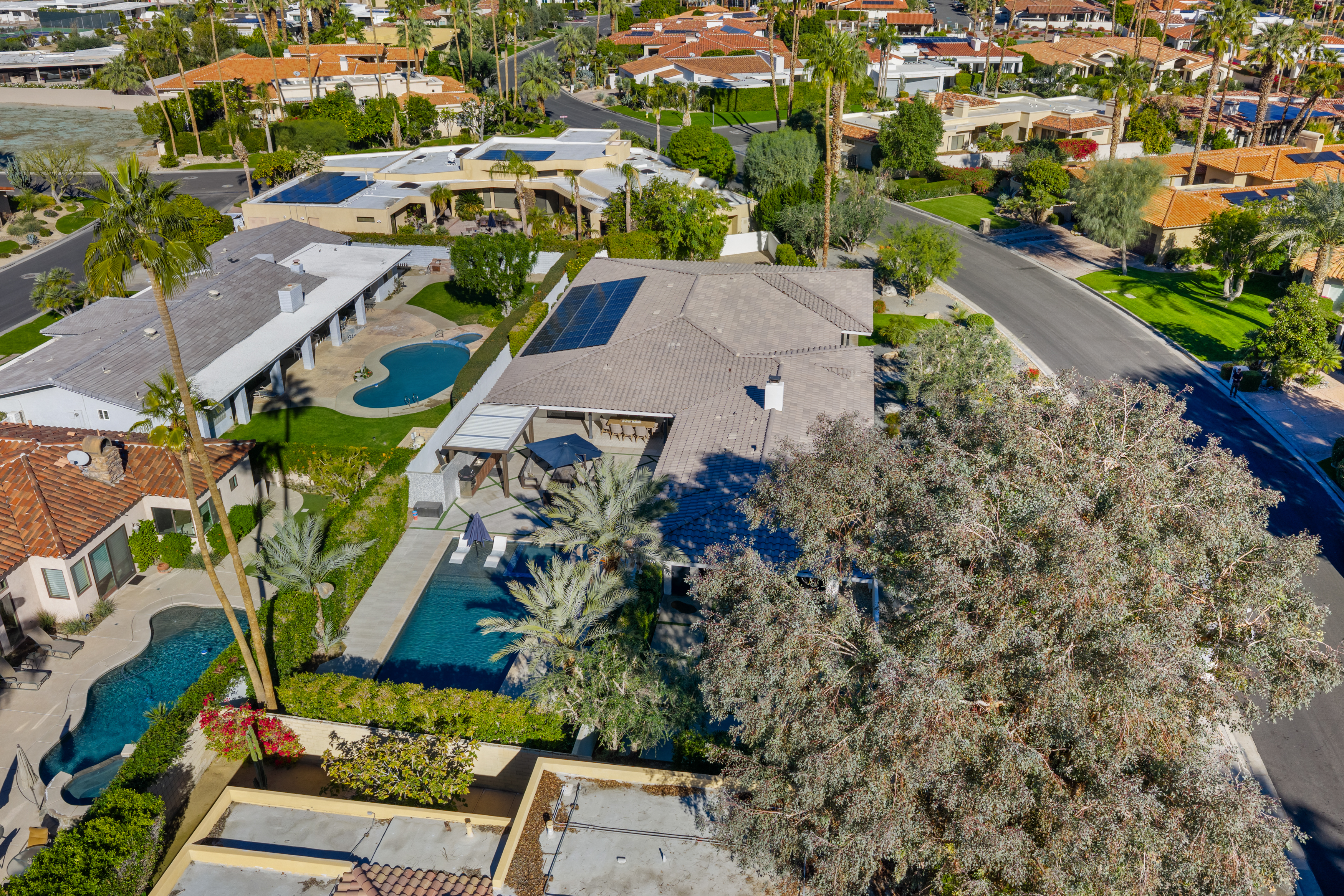 This is an aerial view of a residential property showcasing lush landscaping, including palm trees and manicured lawns. Key features include a swimming pool and multiple houses with red tile roofs, typical of a suburban neighborhood. The property appears well-maintained and offers a private outdoor living space.