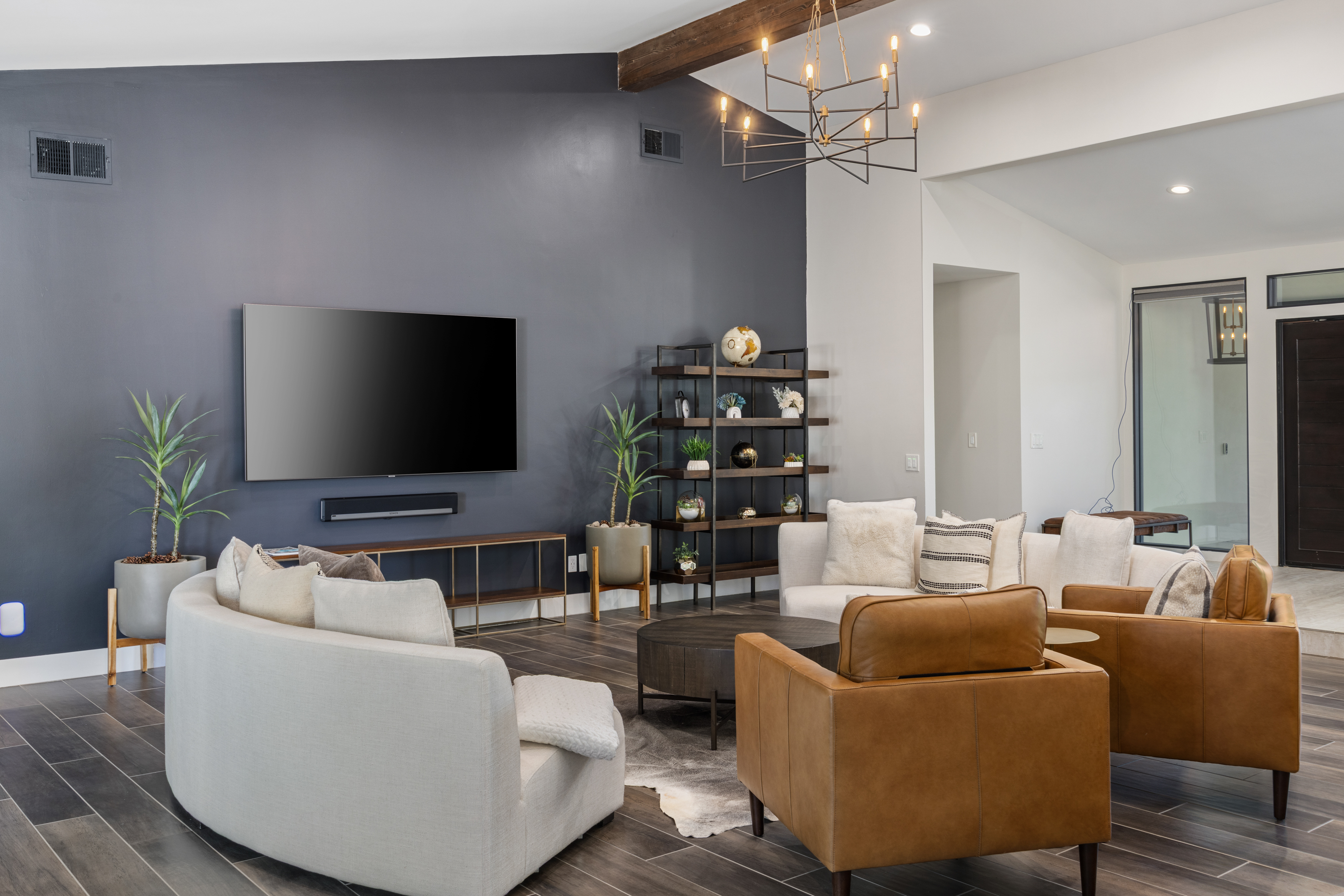 This is an interior shot of a modern living room featuring a contemporary design. A large dark gray wall serves as the backdrop for a large screen television. The room is furnished with a curved sofa, light-colored armchairs, a round coffee table, and stylish shelving, exemplifying comfortable and modern living.
