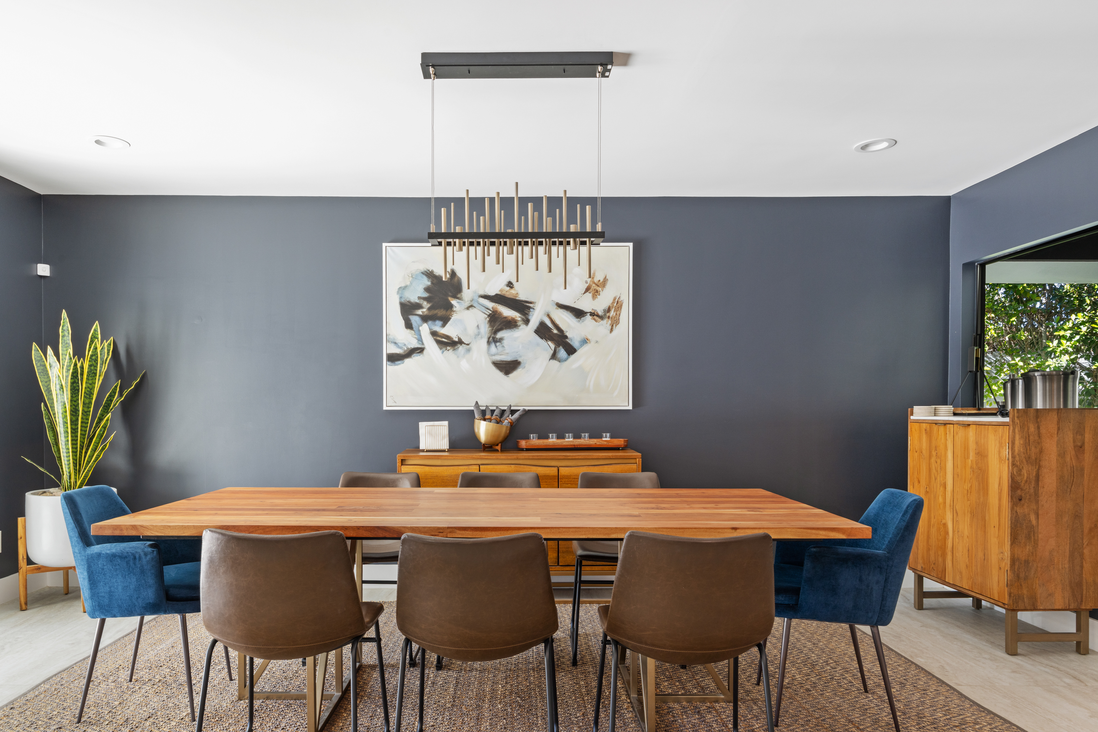 This is an interior shot of a dining room. The room features a long wooden dining table surrounded by brown and blue upholstered chairs. An abstract art piece hangs above a wooden console against a dark grey wall. A striking, modern chandelier hangs above the table creating an elegant ambiance.