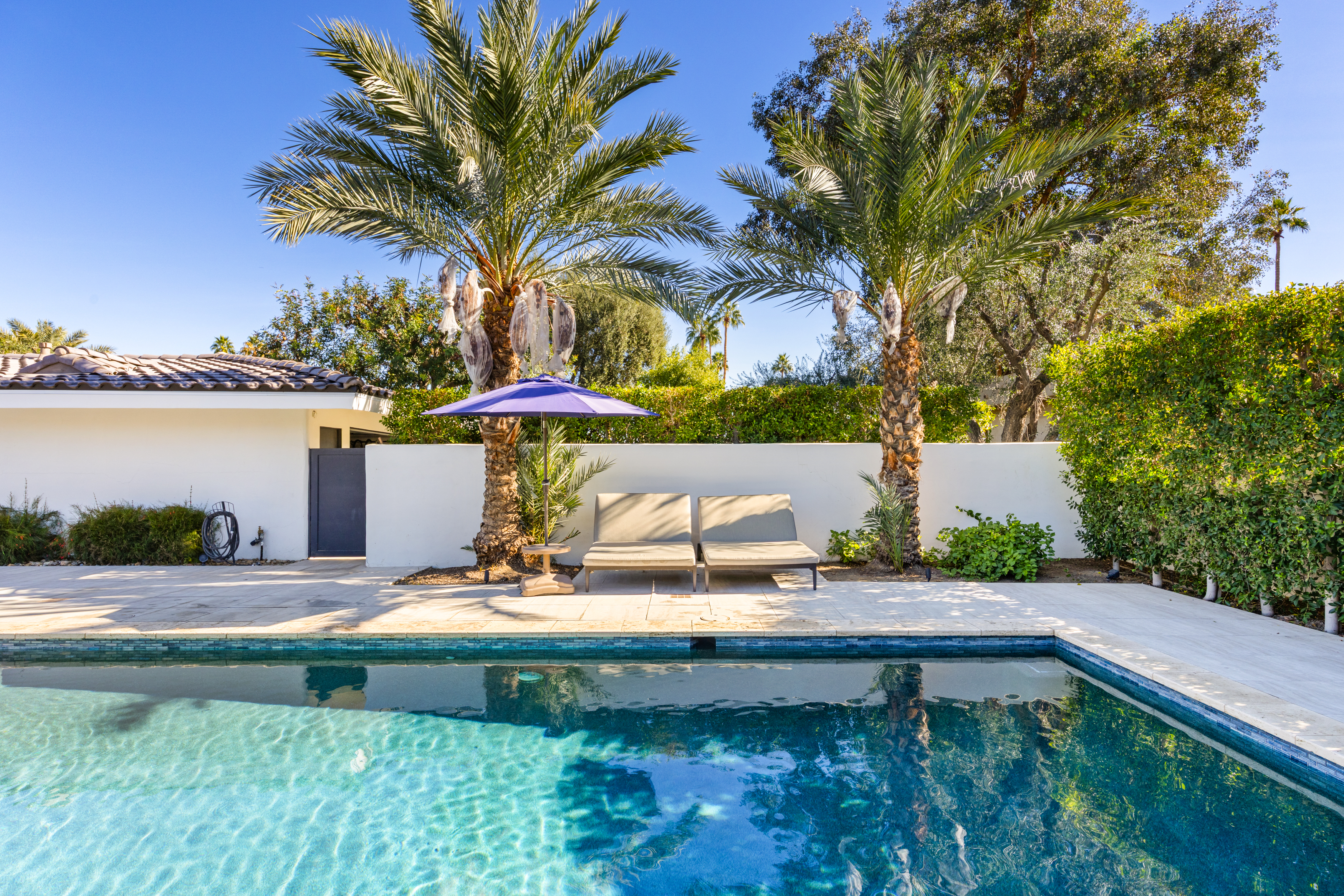 This image showcases a serene pool area, ideal for relaxation and entertainment. Two lounge chairs sit invitingly beside the pool, shaded by palm trees and a blue umbrella. The turquoise water glistens in the sunlight, creating a refreshing and luxurious ambiance.