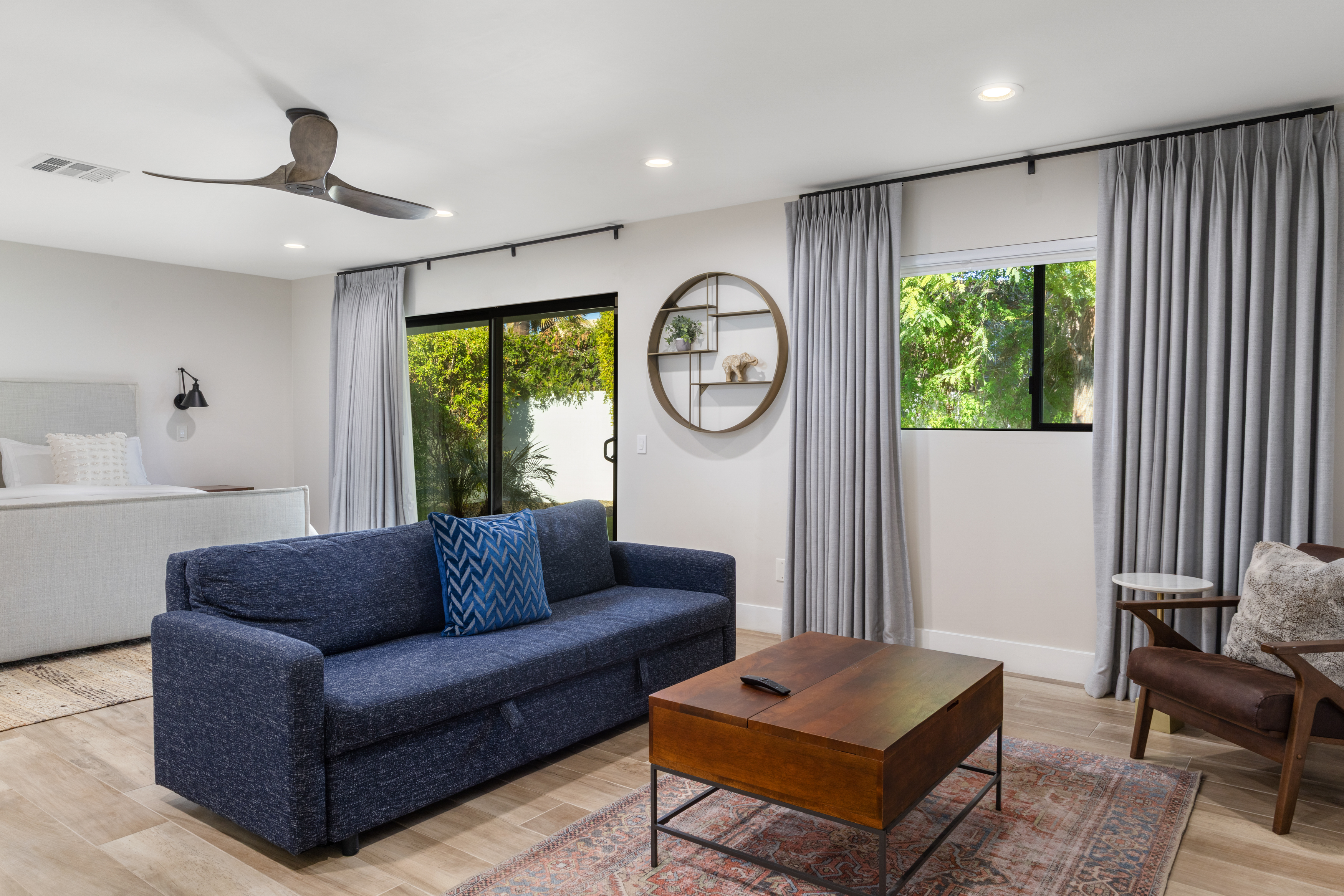 This interior shot showcases a well-lit living space with a contemporary design. The room features a blue sofa with a matching decorative pillow, a wooden coffee table placed on a patterned rug, and a stylish armchair with a side table. Large windows with gray curtains filter natural light, contributing to a warm and inviting atmosphere.