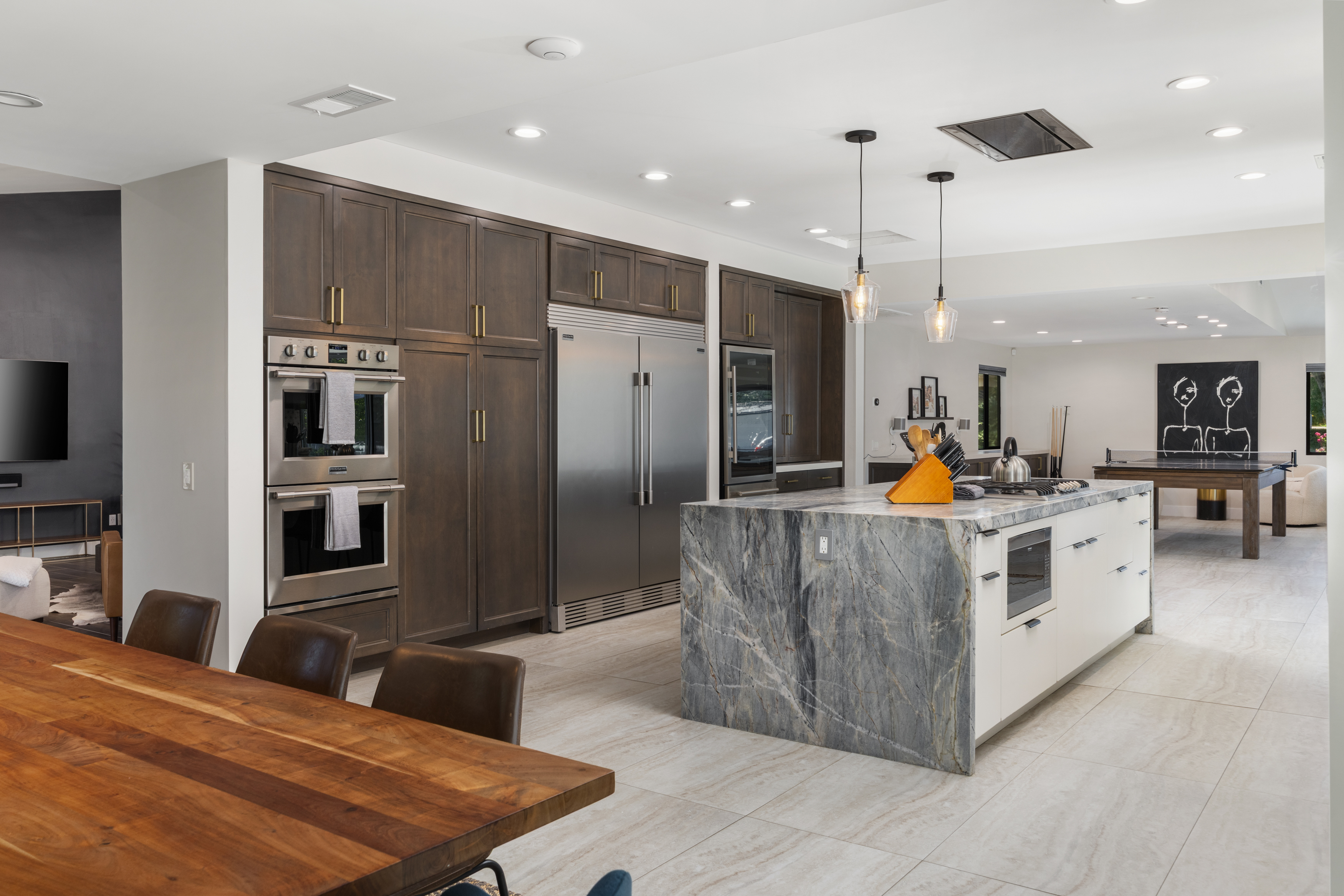 This interior shot showcases a modern kitchen with dark wood cabinetry and stainless steel appliances, including a double oven and refrigerator. A large island with a marble countertop is the focal point, complemented by pendant lighting. The open floor plan connects the kitchen to a dining area with a wooden table and chairs, as well as a living area with a ping pong table.