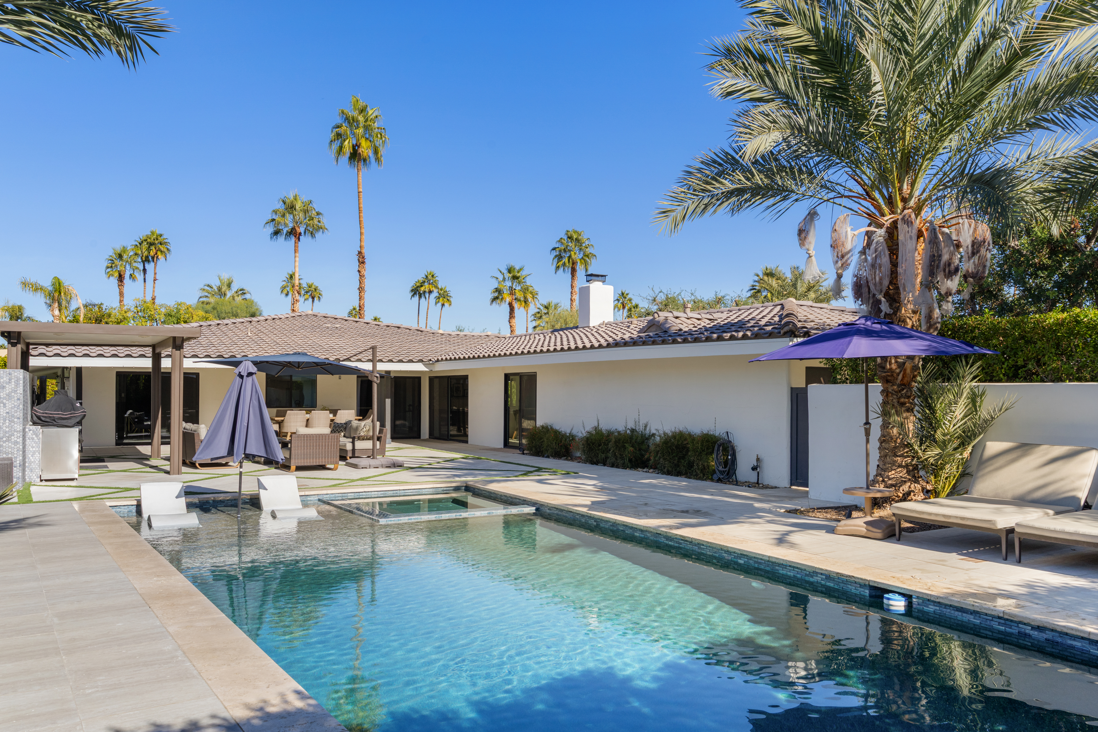 This image showcases a luxurious backyard pool and spa area, perfect for relaxation and entertainment. The pool's clear blue water invites a refreshing dip, while the spa offers a cozy retreat. The surrounding patio features comfortable lounge furniture and a shaded dining area, complementing the modern architecture of the home.