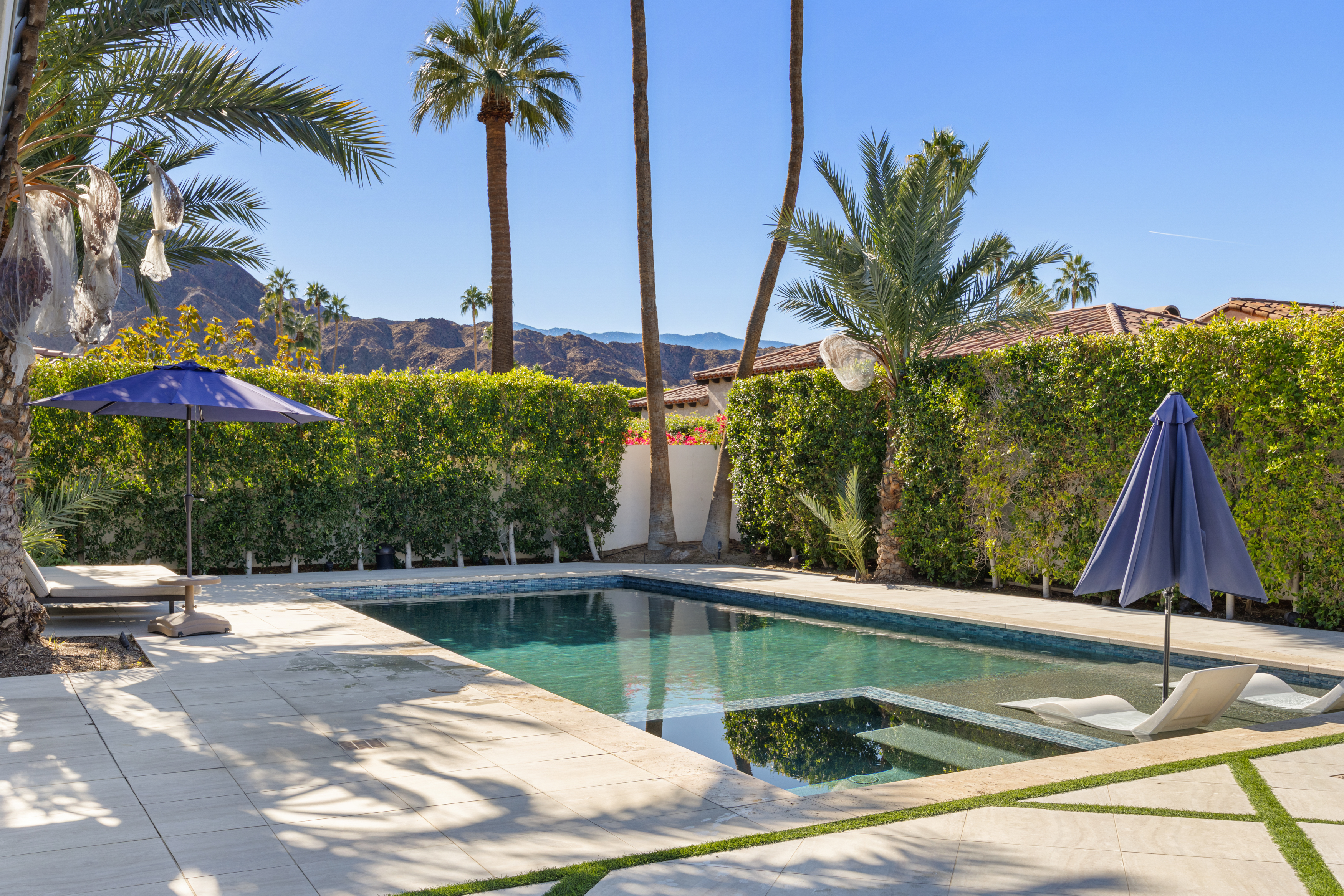 This image showcases a luxurious outdoor swimming pool area. The pool features a modern design with integrated lounge chairs, surrounded by neatly paved decking and lush green manicured hedges. Tall palm trees and views of distant mountains create a resort-like ambiance that would be highly appealing to potential buyers.