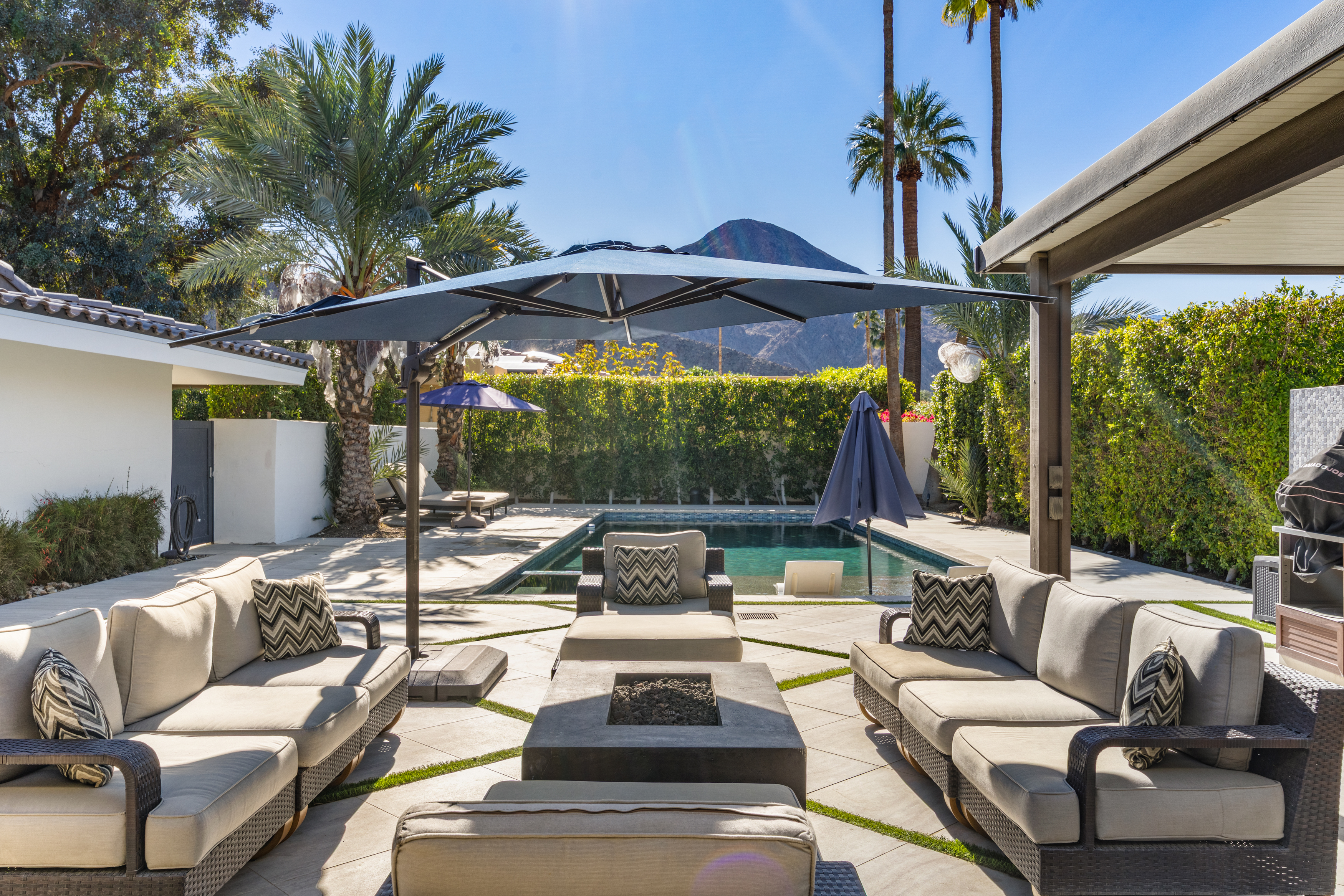 This exterior shot showcases a luxurious pool area, complete with stylish outdoor furniture arranged around a modern fire pit. A large patio umbrella provides shade, while mature landscaping and palm trees enhance the resort-like ambiance. In the background, a mountain rises, adding a scenic element to the backyard oasis.
