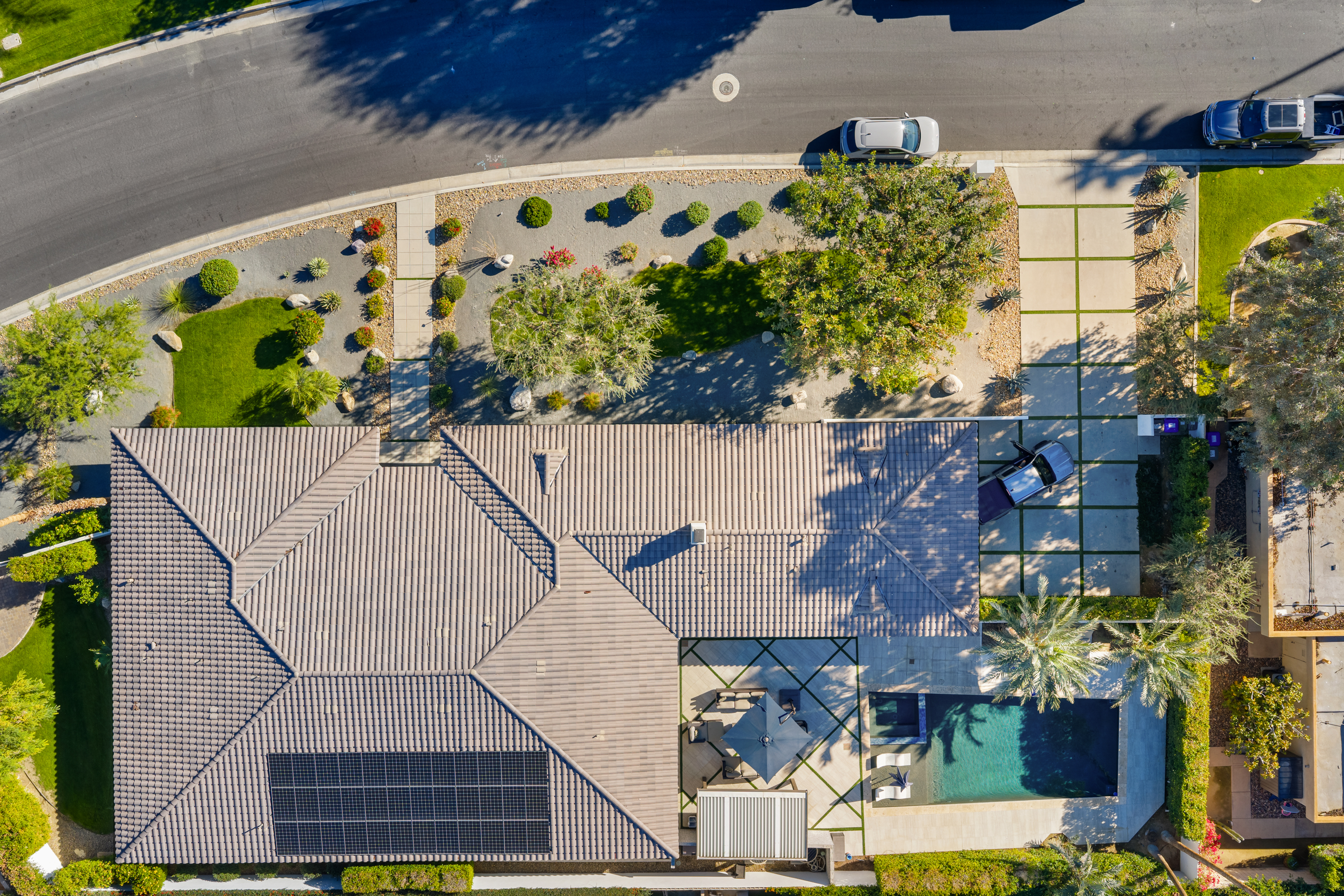 This aerial shot showcases a well-maintained property featuring a tiled roof with solar panels, a sparkling pool with lounge seating, and lush landscaping. The layout emphasizes outdoor living with a covered patio and manicured grounds. The property boasts a modern design with clean lines and thoughtful hardscaping, enhancing its curb appeal.