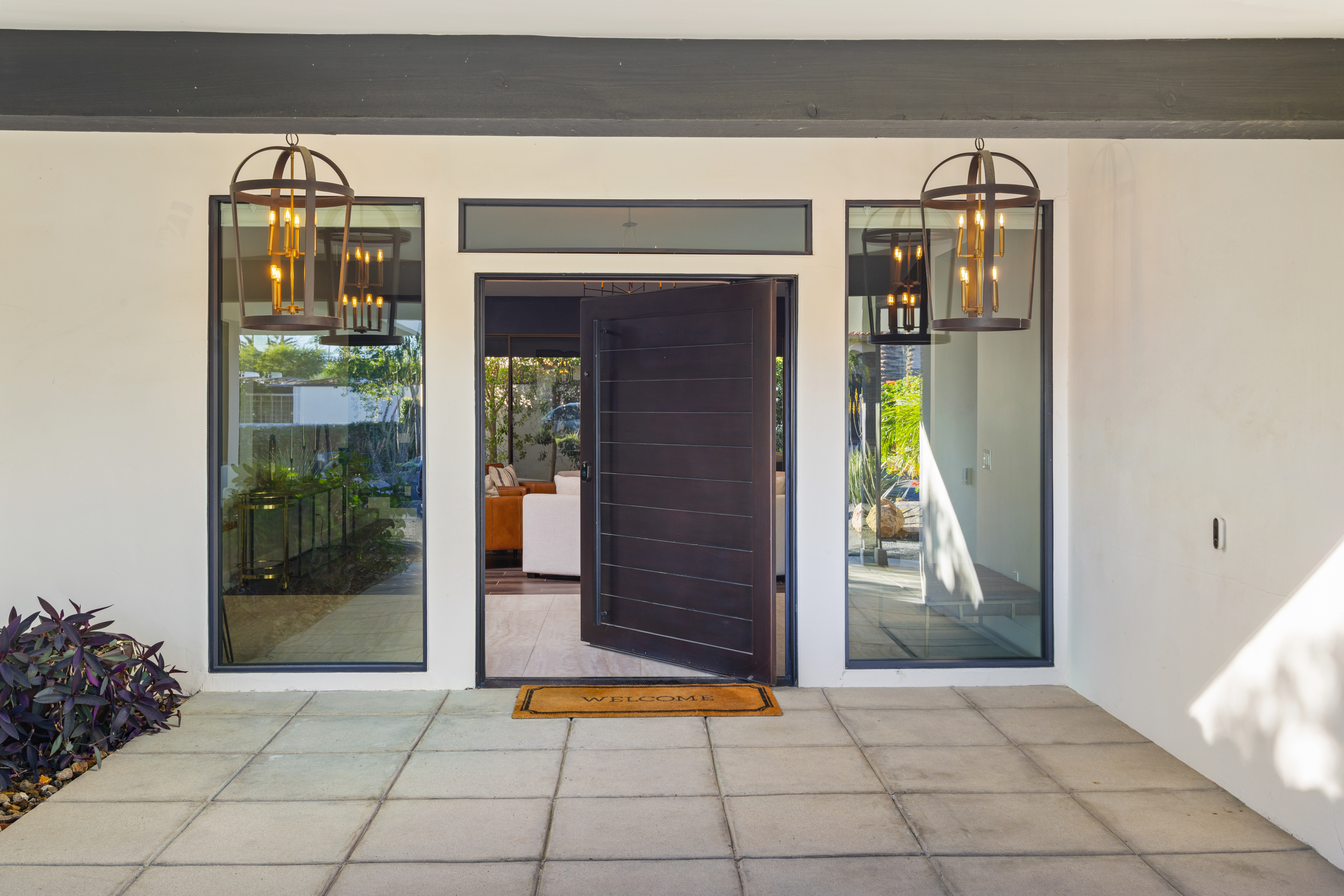 This is an inviting entryway to a contemporary home. A dark wood door is framed by sidelight windows and a transom window, with stylish hanging light fixtures on either side. A 'WELCOME' mat sits on the tiled patio, adding a warm touch.