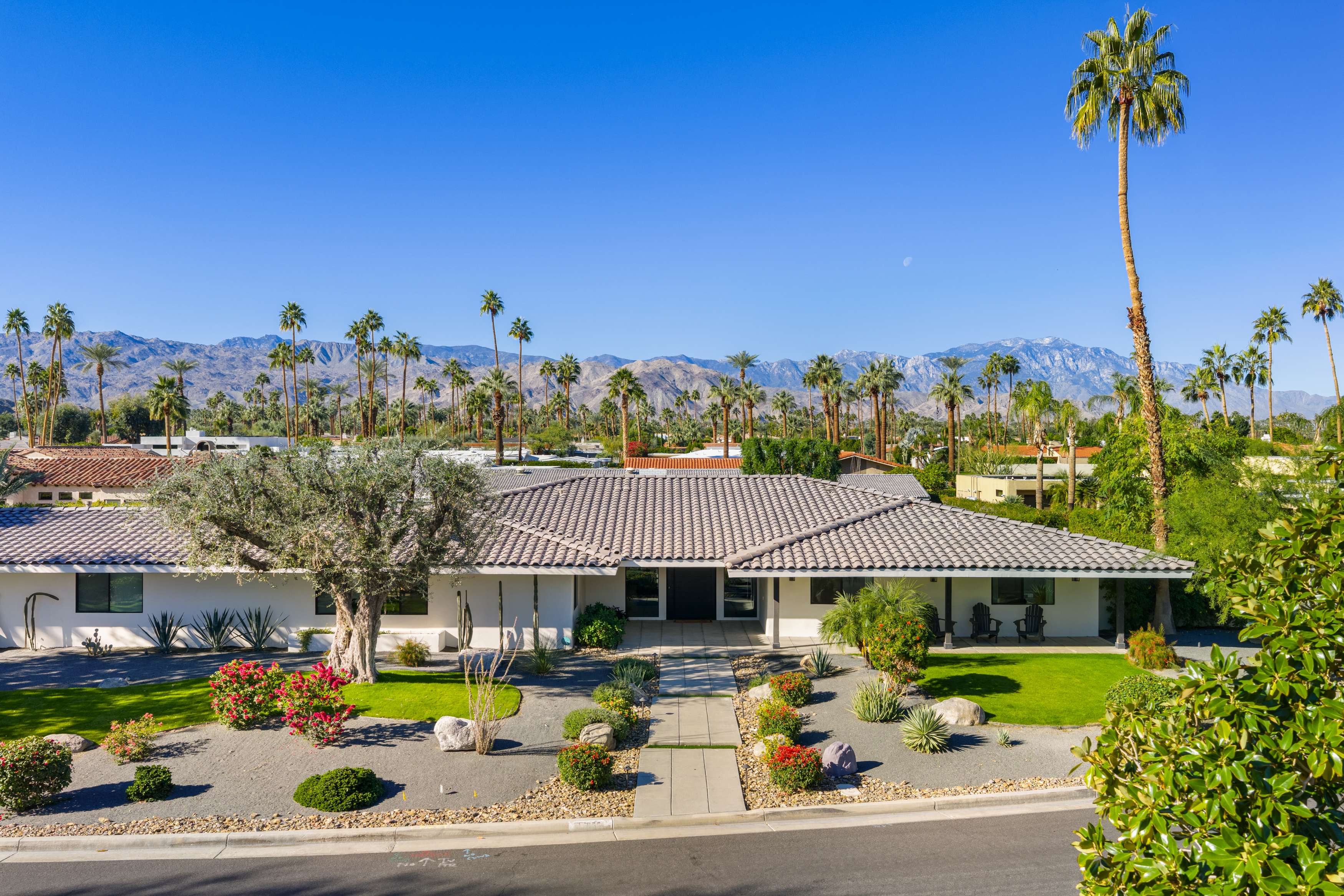 This is a front exterior view of a well-maintained, single-story home in what appears to be Palm Springs, California. The house features a tan/grey tiled roof, a white facade, and a desert-landscaped front yard with gravel, greenery and flowers. Mountains and palm trees are prominent in the background creating a classic California vista.