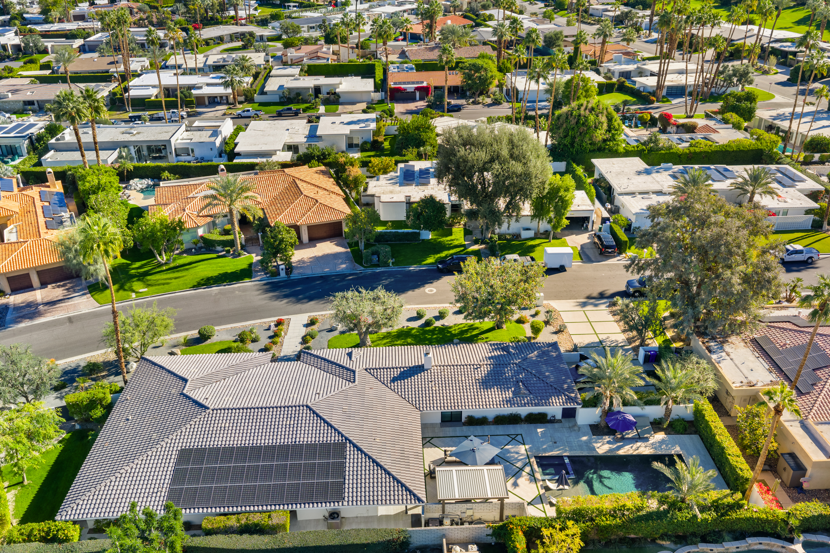 This aerial view showcases a well-maintained residential neighborhood, highlighting the lush landscaping and variety of architectural styles. The home in the foreground features a grey tiled roof with solar panels and a private swimming pool flanked by manicured greenery. The overall impression is one of upscale suburban living with attention to detail and outdoor amenities.