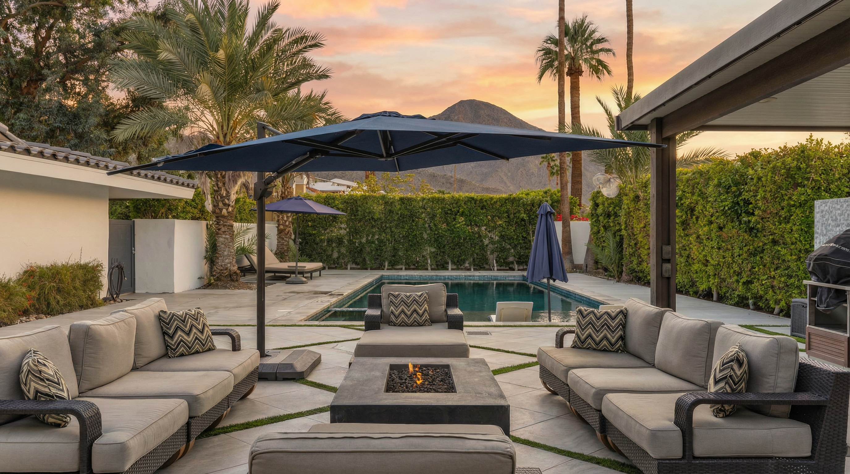 This exterior shot features a beautifully designed pool area with stylish outdoor seating, including sofas and lounge chairs around a fire pit. The pool's pristine water and well-maintained patio are enhanced by lush green hedging and palm trees, providing a luxurious desert oasis. An umbrella offers shade for the seating arrangements.