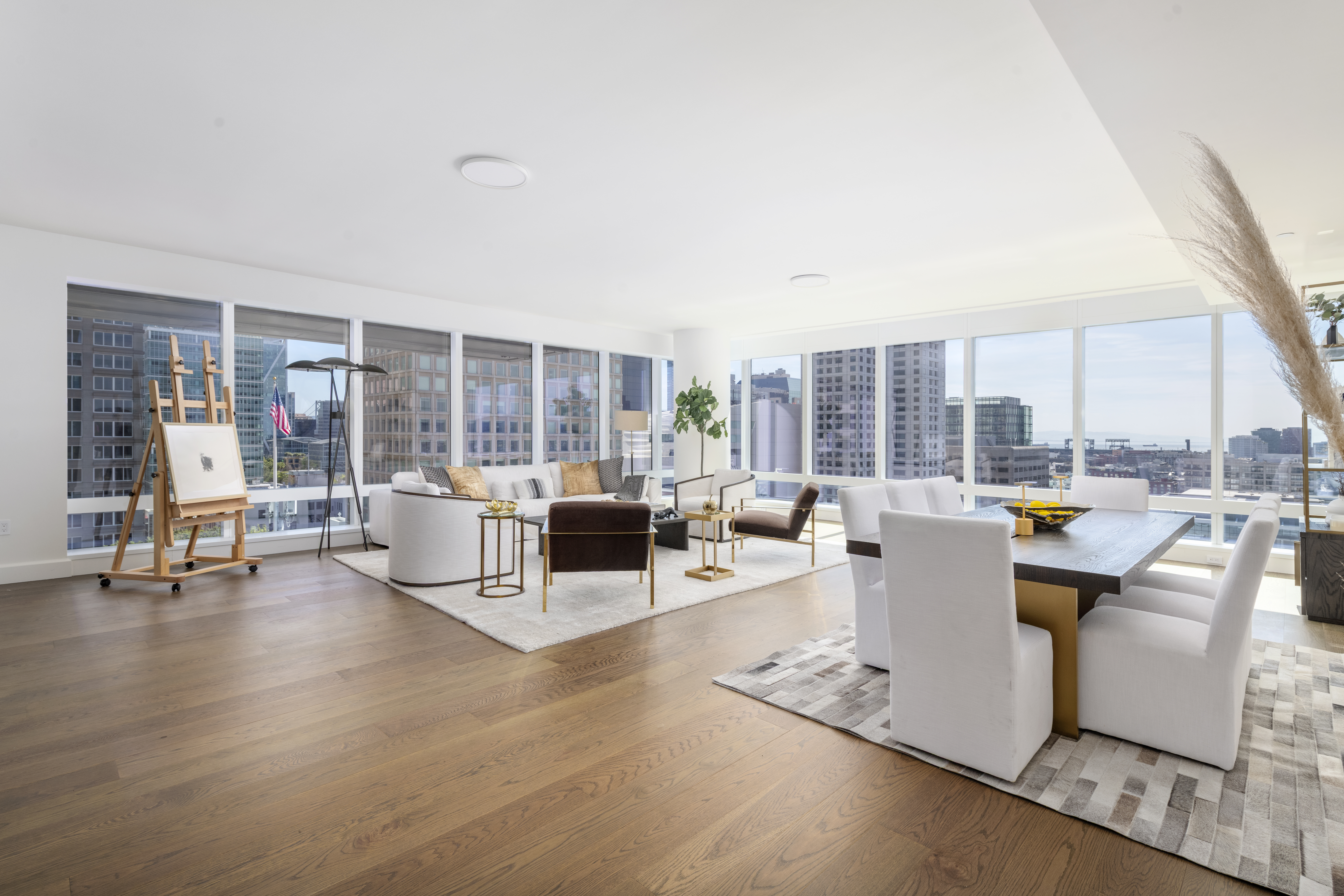 This expansive, open-concept living and dining area features light wood flooring and floor-to-ceiling windows that provide panoramic city views. The living space is anchored by a curved sofa, a dark armchair, and a central coffee table, while the adjacent dining area includes a large table with white chairs set upon a textured rug. The room is bright and airy, showcasing a sophisticated, modern aesthetic with high-end furnishings and an artist's easel near the window.