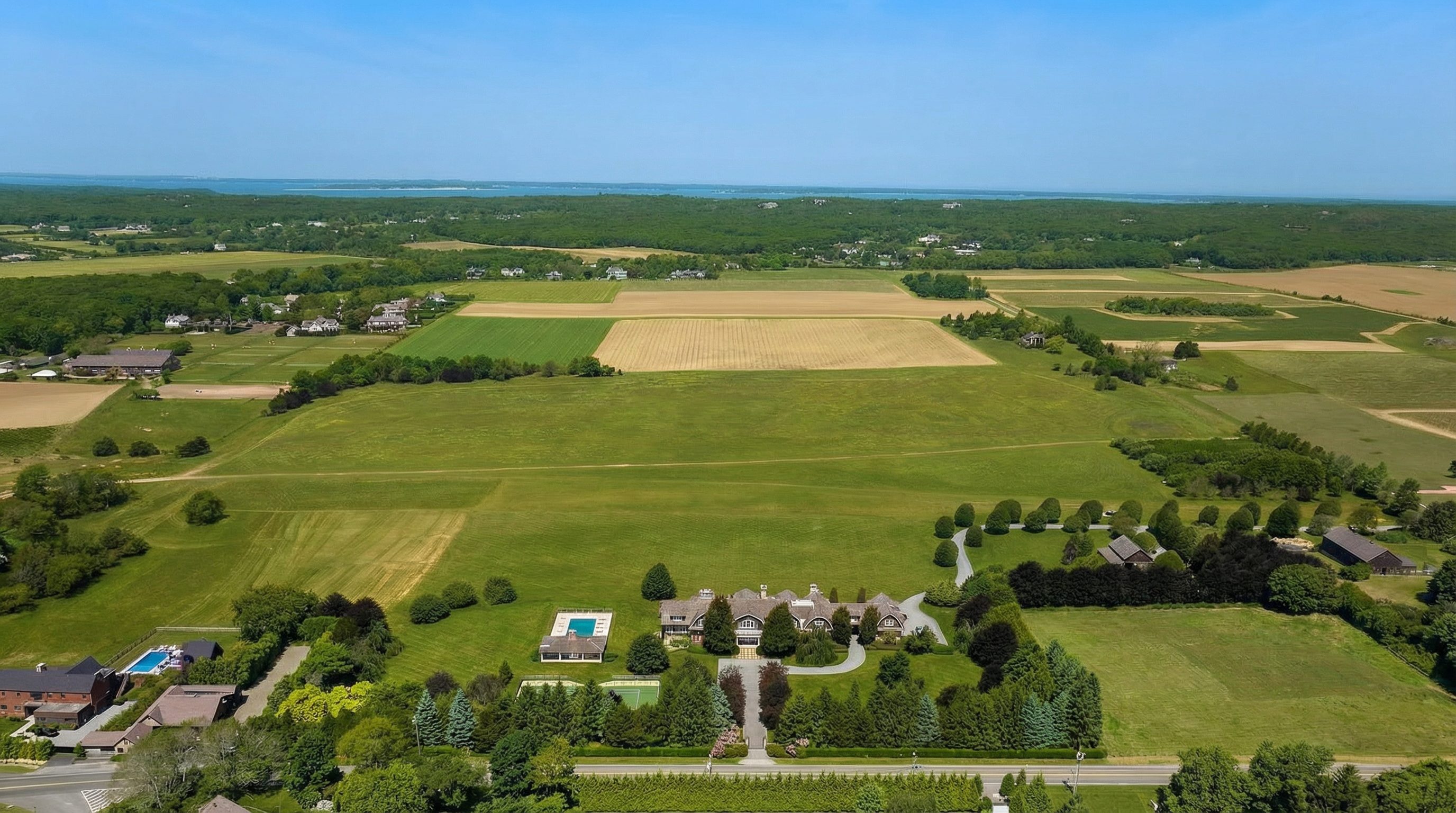 This is an aerial view of a sprawling estate featuring lush green lawns, mature trees, and agricultural fields. The main residence is centrally located, complete with a swimming pool, tennis court and well-manicured grounds. The property's vast size and meticulously maintained landscaping evoke a sense of privacy and luxury.