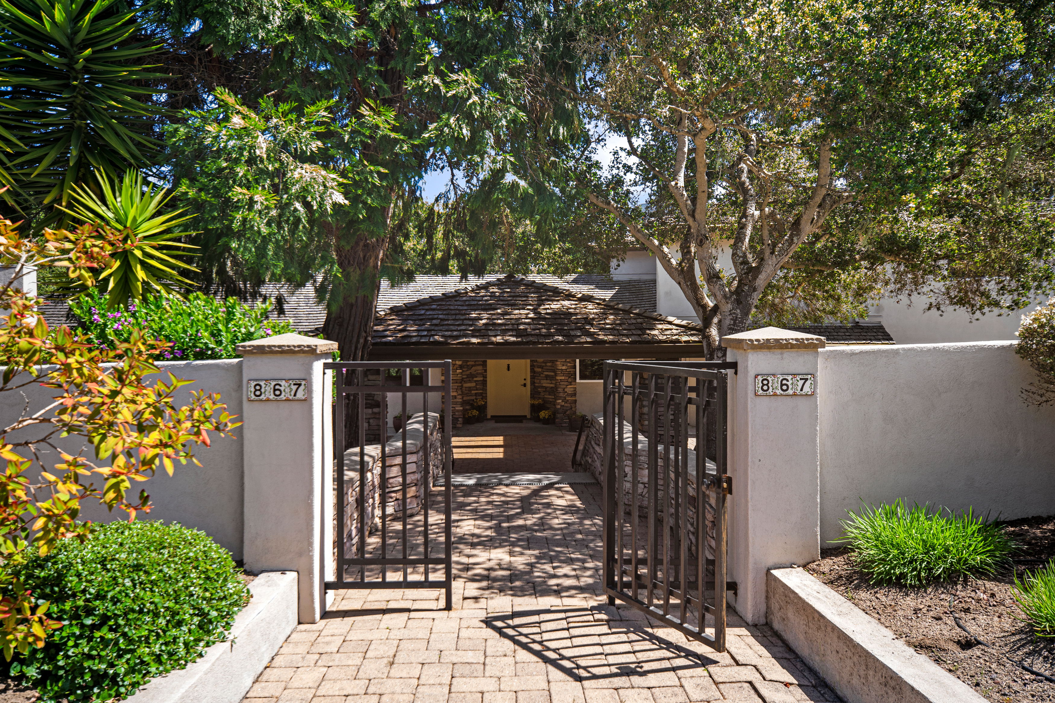 This inviting entryway features a gated brick-paved path leading toward a front door framed by stone masonry. Mature trees provide dappled shade over the walkway, creating a serene and private atmosphere. The property is clearly marked with the number 867 on both stone pillars flanking the entrance.