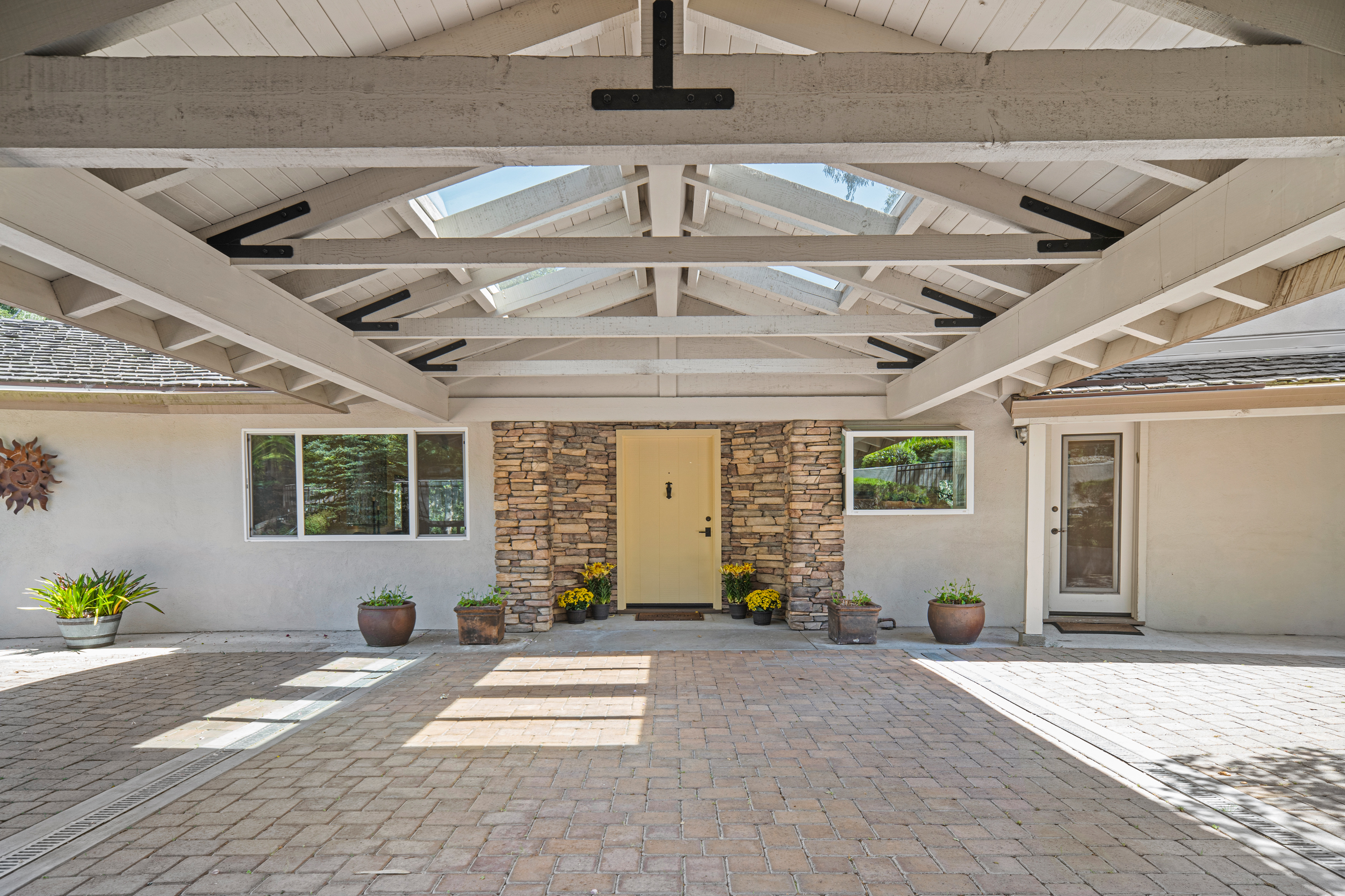 This inviting entryway features a covered porte-cochère with exposed wooden beams and a skylight, providing a bright and sheltered approach to the home. The front door is painted a soft yellow and is framed by a textured stone surround, flanked by potted plants and yellow flowers. The ground is paved with light-toned brick, creating a cohesive and well-maintained arrival area.