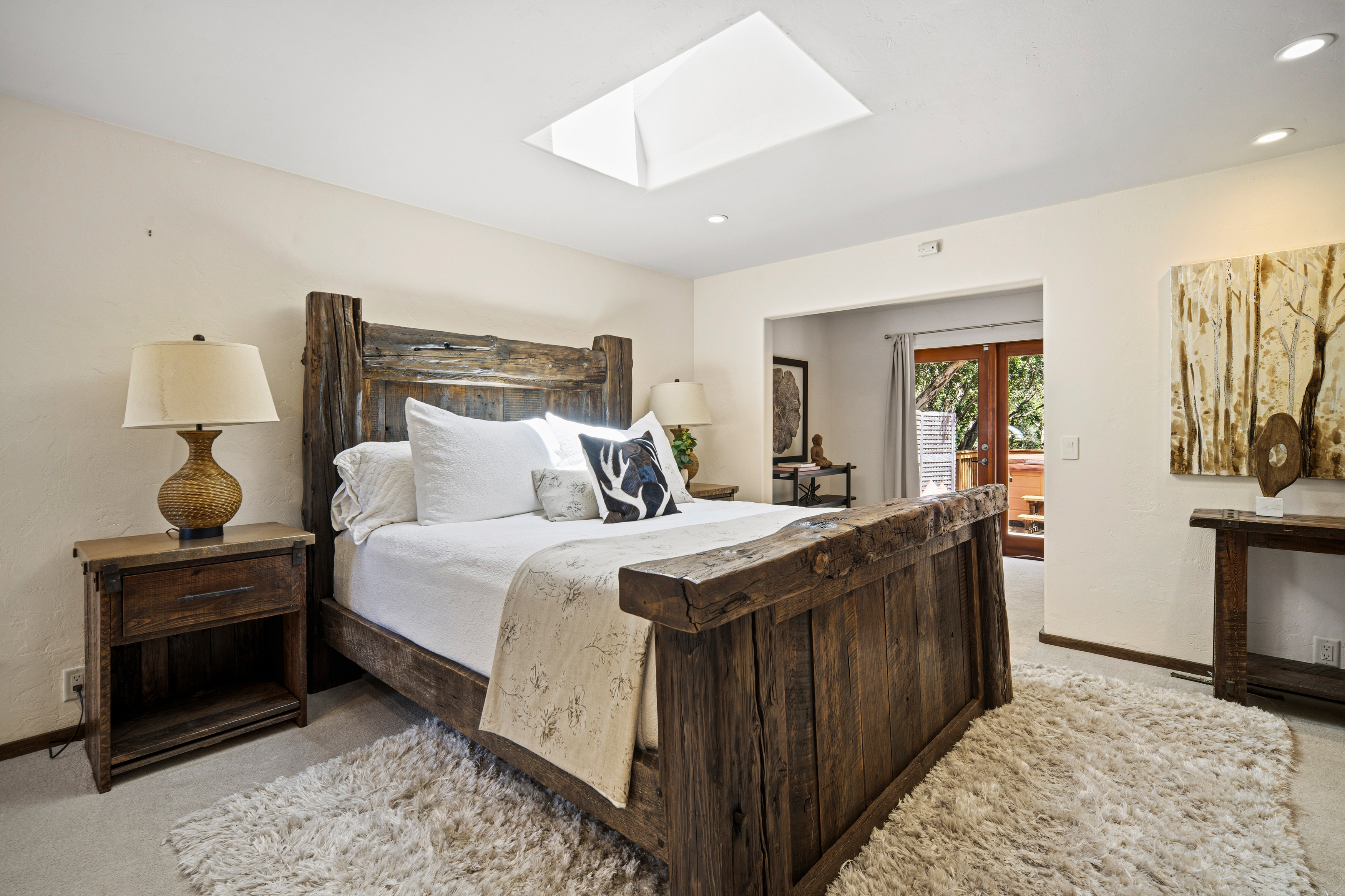 This primary bedroom features a rustic aesthetic centered around a substantial, dark-stained wood bed frame with a prominent headboard and footboard. The room is illuminated by a large skylight and a pair of bedside lamps, creating a warm and inviting atmosphere. A doorway leads to an adjacent space with a view of an outdoor deck, while a textured area rug adds comfort to the neutral-toned space.