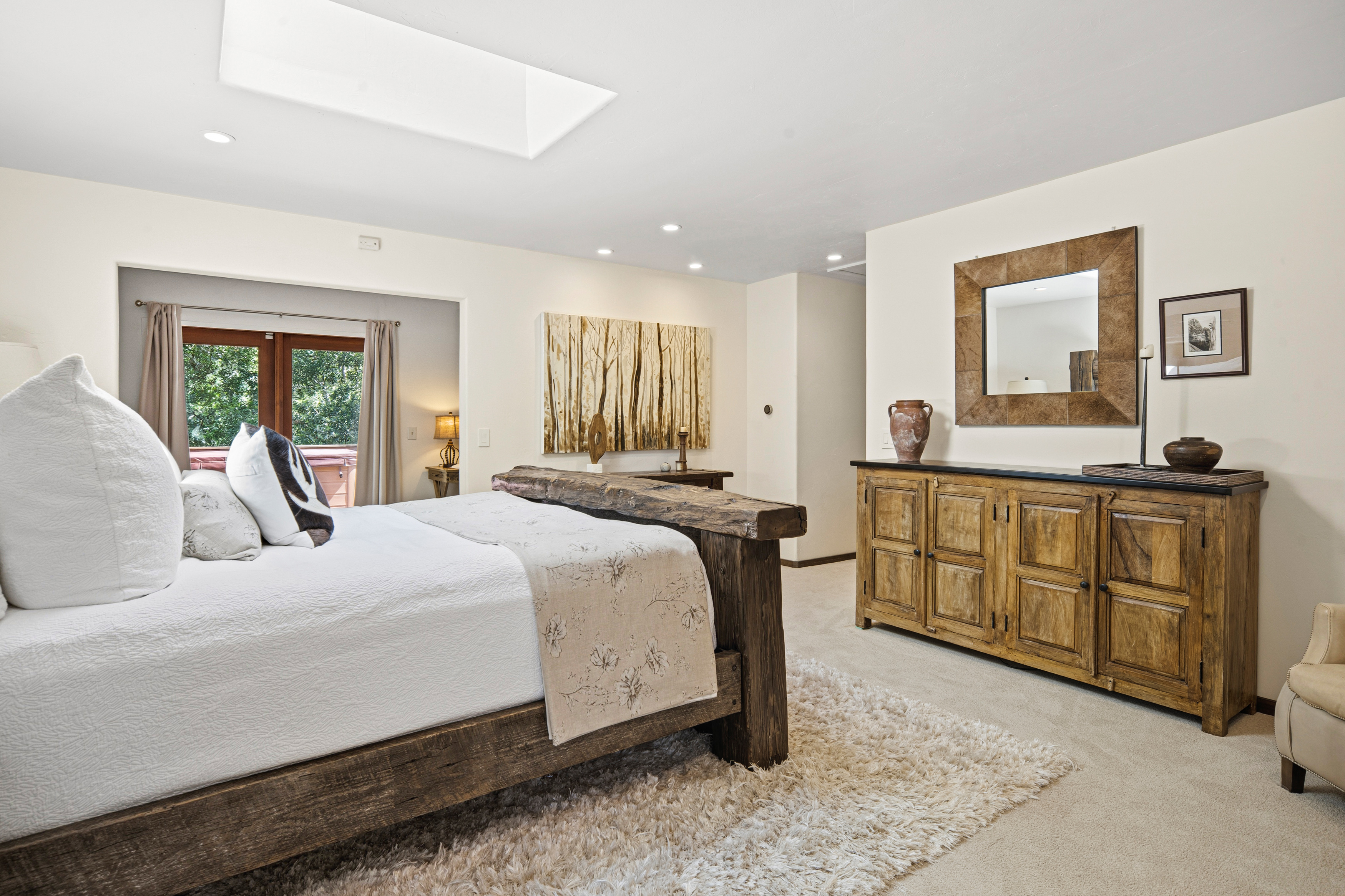 This primary bedroom features a rustic aesthetic with a substantial wood-frame bed as the focal point, complemented by a textured, light-colored area rug. A large wooden dresser with a matching framed mirror sits against the wall, while a skylight overhead provides ample natural light. The room opens into an adjacent space with a view of an outdoor deck, creating a bright and inviting atmosphere.