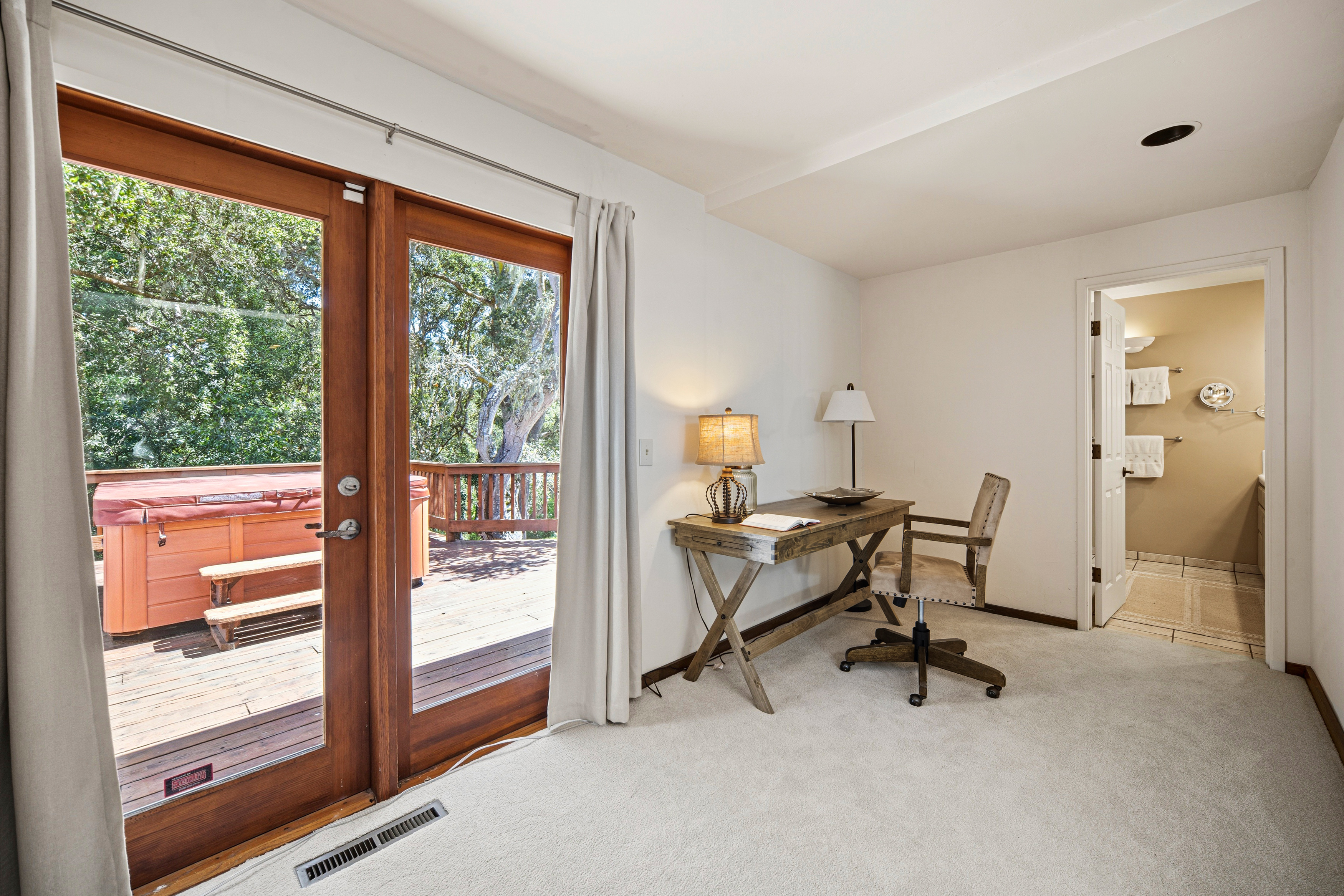 This bright home office features neutral carpeting and white walls, centered around a rustic wooden desk with a matching chair. Large glass doors provide a seamless transition to an outdoor deck, where a hot tub is visible, creating a relaxing indoor-outdoor connection. The room also includes a doorway leading into an adjacent bathroom, offering convenience and a functional layout.