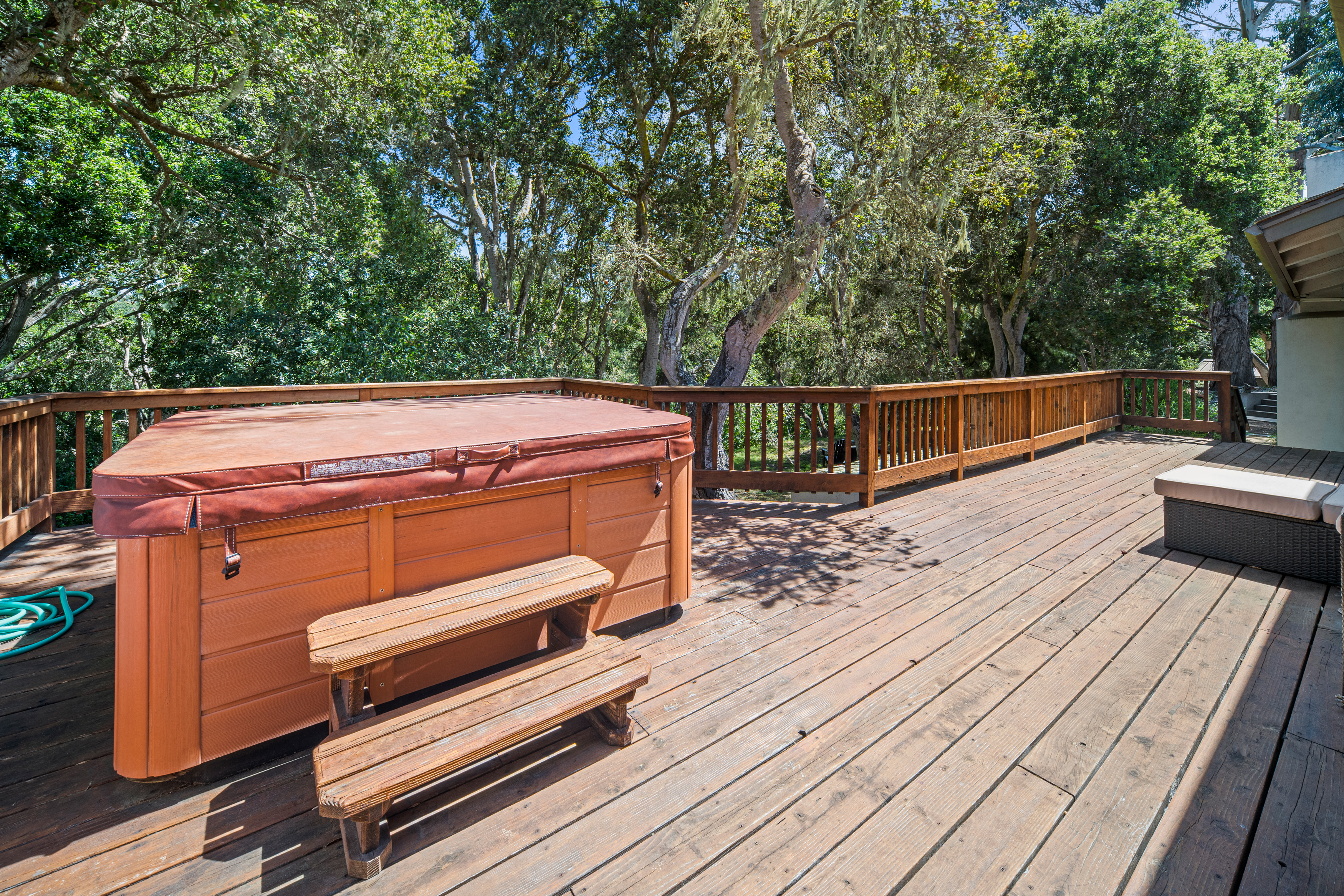This spacious wooden deck features a large hot tub with a protective cover and matching wooden steps, set against a backdrop of lush, mature trees. The deck is enclosed by a wooden railing and includes a cushioned outdoor seating area to the right, creating a private and relaxing outdoor retreat. The perspective is from a slightly elevated angle, capturing the expansive wood plank flooring and the serene, wooded surroundings.