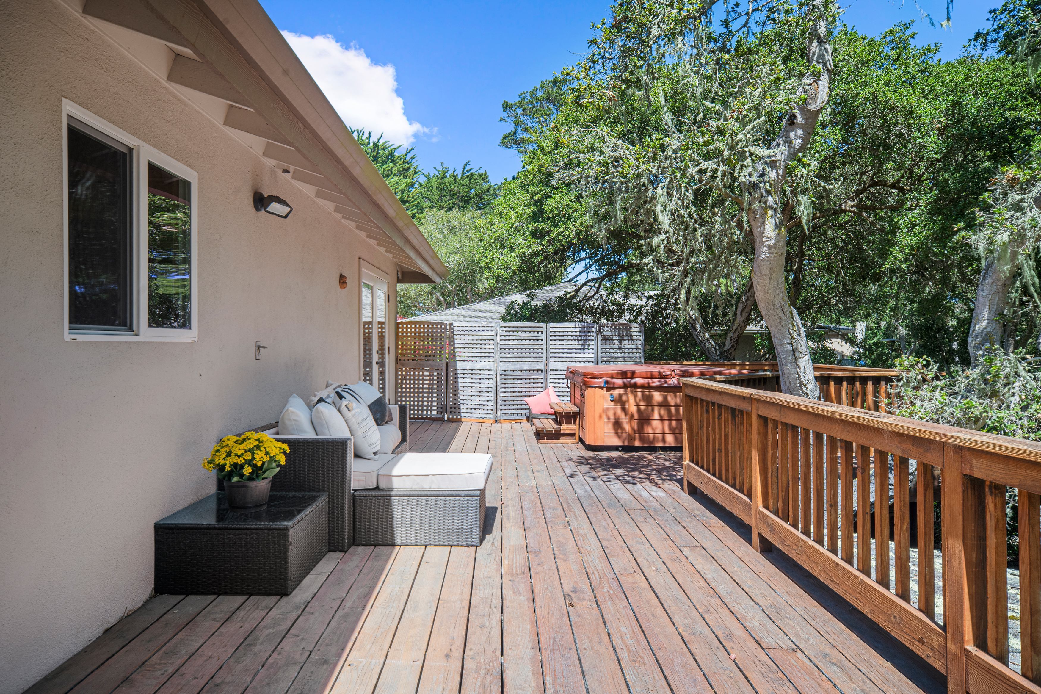 This spacious wooden deck features a comfortable outdoor seating area with grey wicker furniture and a potted plant, perfect for relaxation. A hot tub is positioned near the wooden railing, which overlooks a lush, tree-filled backyard. The scene is captured from a wide-angle perspective, highlighting the seamless transition between the home's exterior and the private, natural outdoor living space.