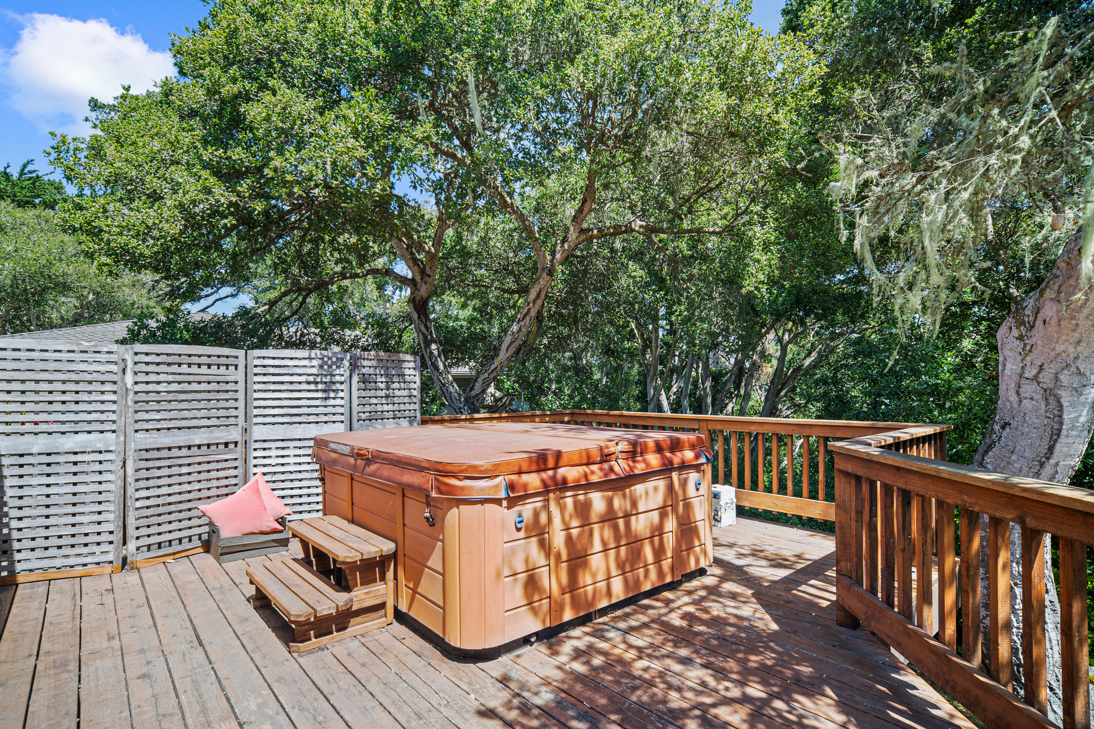 This elevated wooden deck features a large, covered hot tub as its central focal point, complete with a matching set of wooden steps. The space is enclosed by a wooden railing and a privacy screen, offering a secluded outdoor retreat surrounded by mature, leafy trees. The perspective is a wide-angle shot from the deck surface, capturing the inviting and private atmosphere of this outdoor living area.