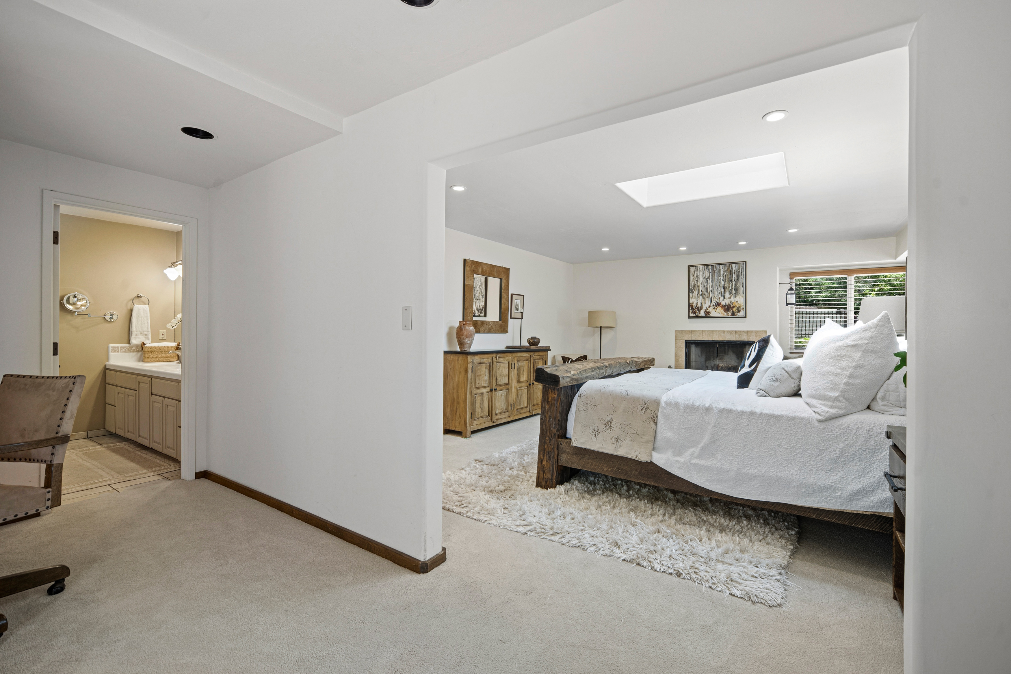 This primary bedroom features a rustic, heavy-timber bed frame as the centerpiece, set against a backdrop of neutral walls and a plush, textured area rug. To the left, an open doorway leads to an en-suite bathroom with a light-wood vanity, while a wooden dresser and a fireplace add warmth to the sleeping area. A skylight overhead provides natural illumination, creating an airy and inviting atmosphere.