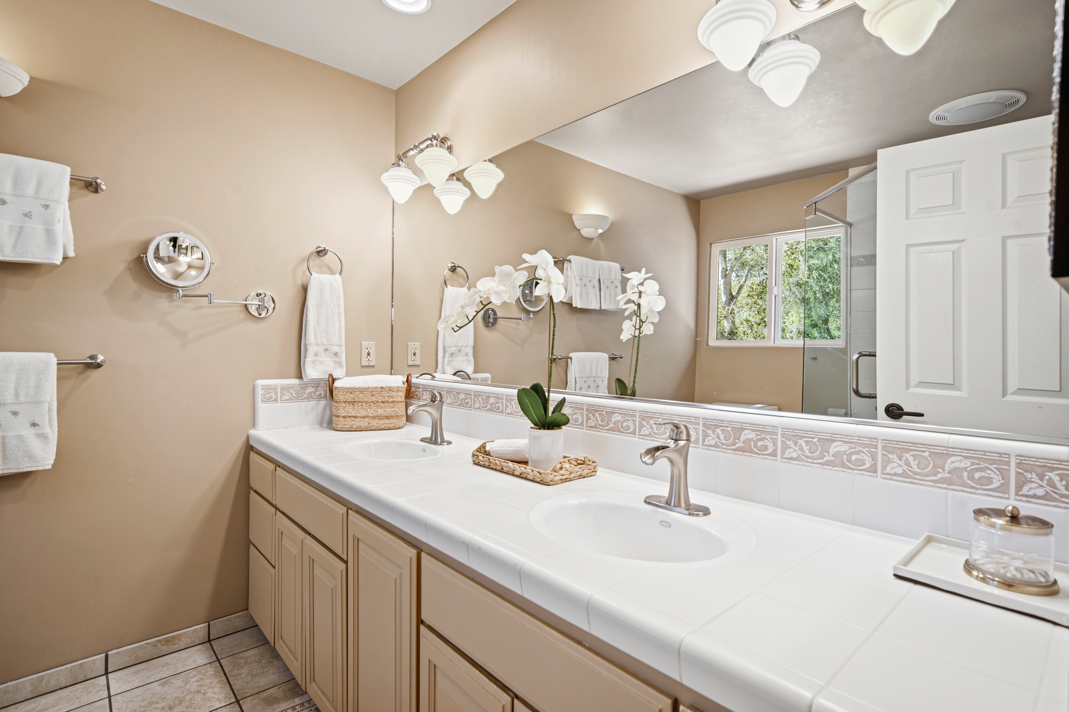 This dual-vanity bathroom features a long, white tiled countertop with two integrated sinks and brushed nickel fixtures. The space is finished with light-toned cabinetry, a large wall-to-wall mirror, and a decorative tile backsplash that adds a touch of texture. A window above the shower area allows natural light to brighten the room, which is accented by a potted orchid and neatly folded towels.