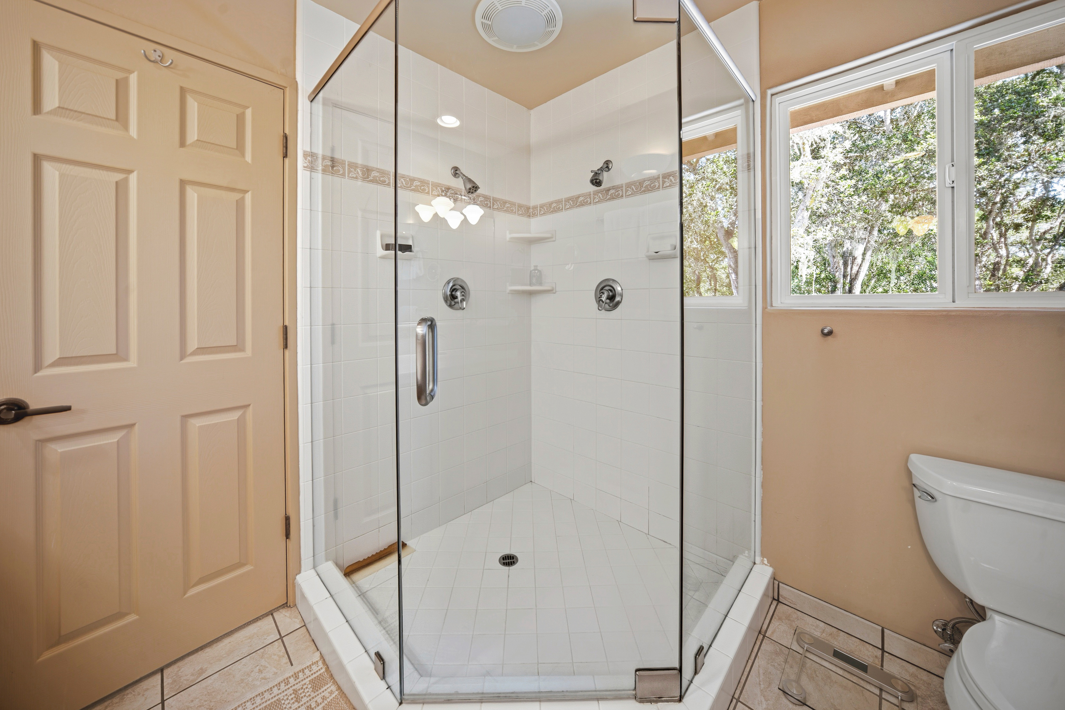 This bathroom features a corner glass-enclosed shower with white tiled walls and a decorative border accent. The space includes a white toilet and a neutral-toned door, with a window providing natural light and a view of the outdoors. The overall aesthetic is functional and bright, highlighting a clean, classic design.