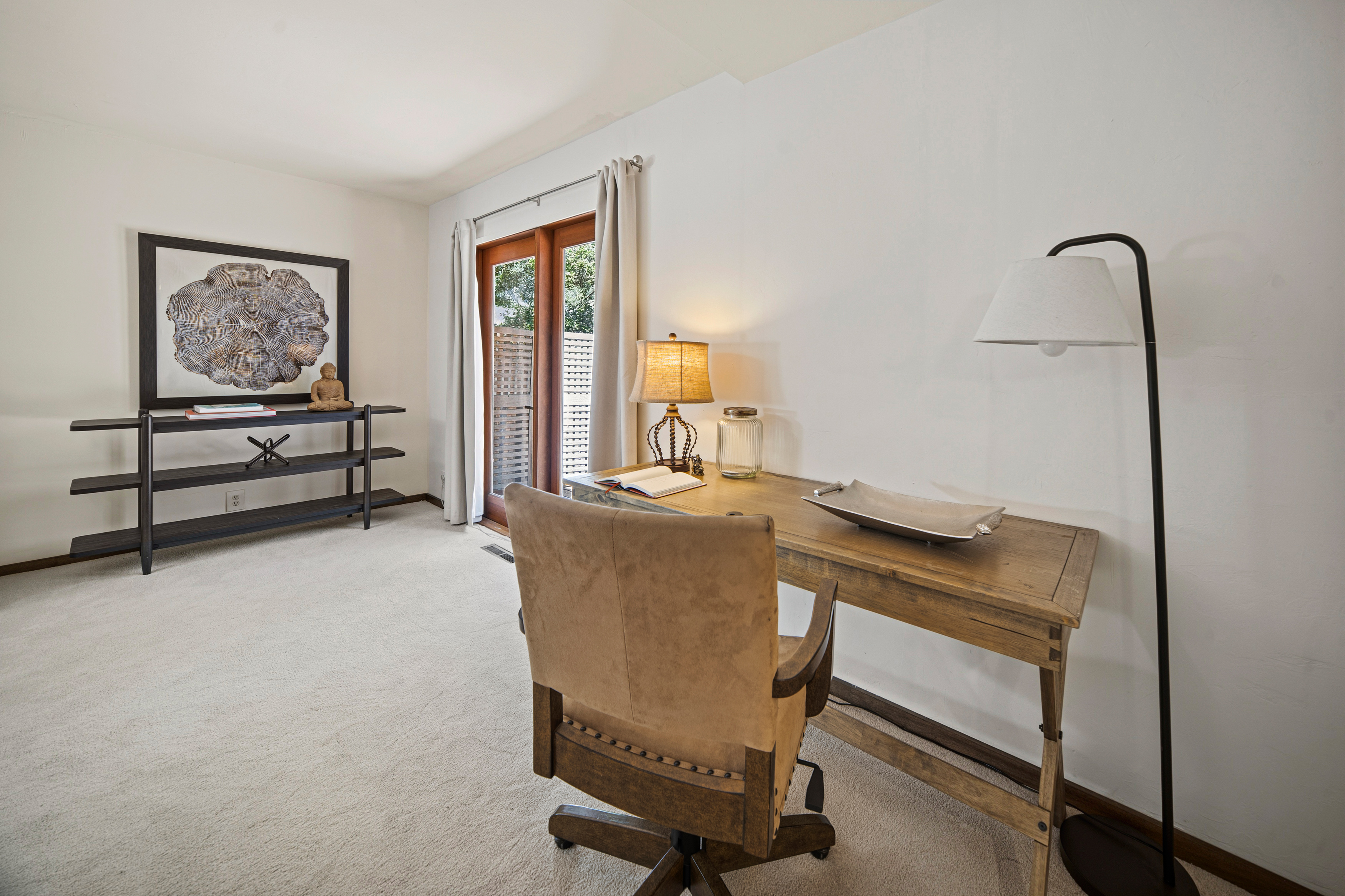 This home office features a rustic wooden desk paired with a leather-upholstered swivel chair, creating a warm and professional workspace. A tall black shelving unit against the left wall displays decorative items, while a floor lamp provides focused lighting near the desk. The room is brightened by a glass-paned door that leads to an outdoor area, framed by simple curtains.