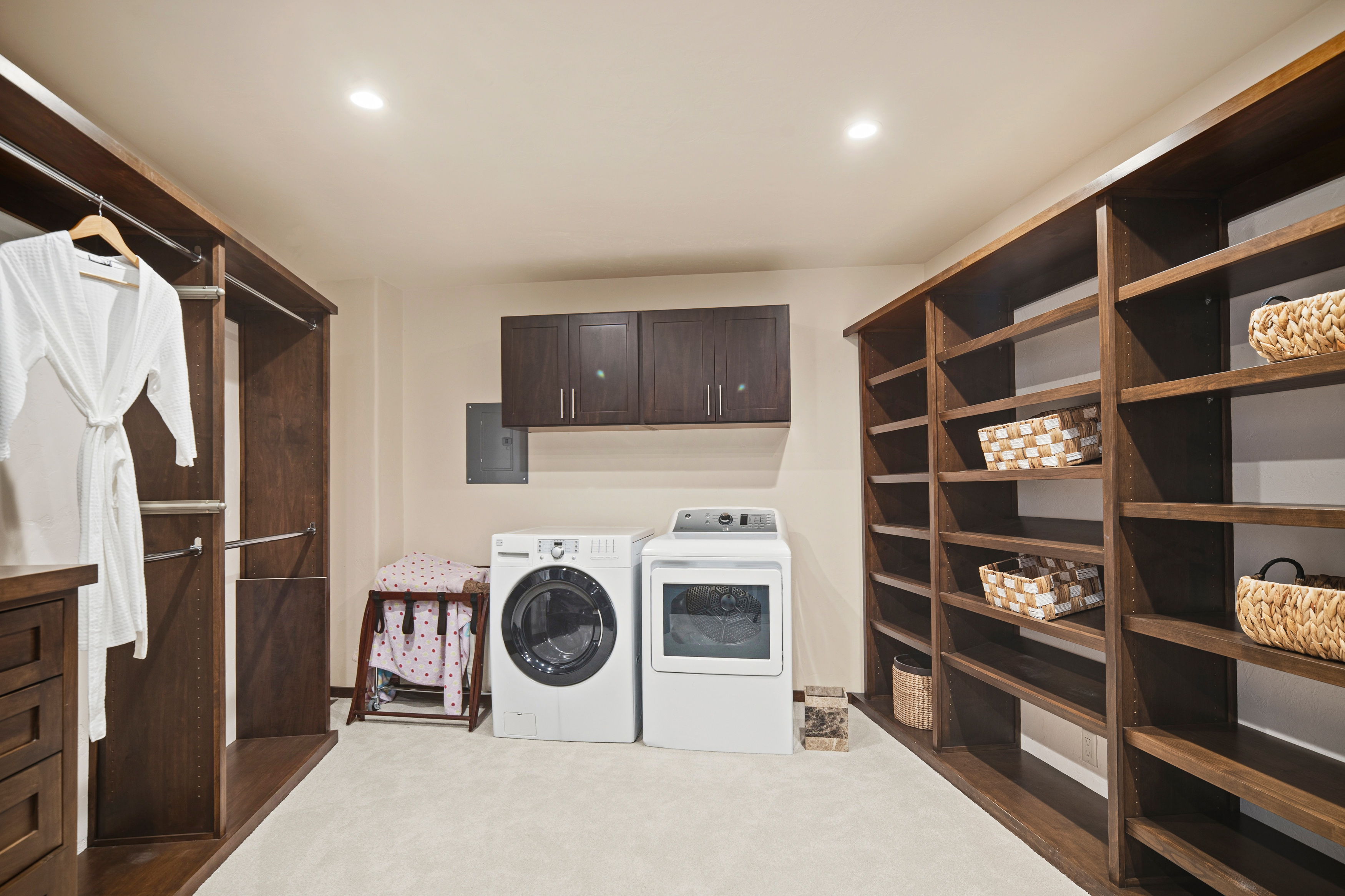 This functional space serves as a combined laundry room and walk-in closet, featuring dark wood cabinetry and shelving units. A white washer and dryer set are positioned centrally beneath wall-mounted cabinets, while open shelving on the right provides ample storage for baskets. The room is finished with light-colored carpeting and recessed lighting, creating a clean and organized utility area.