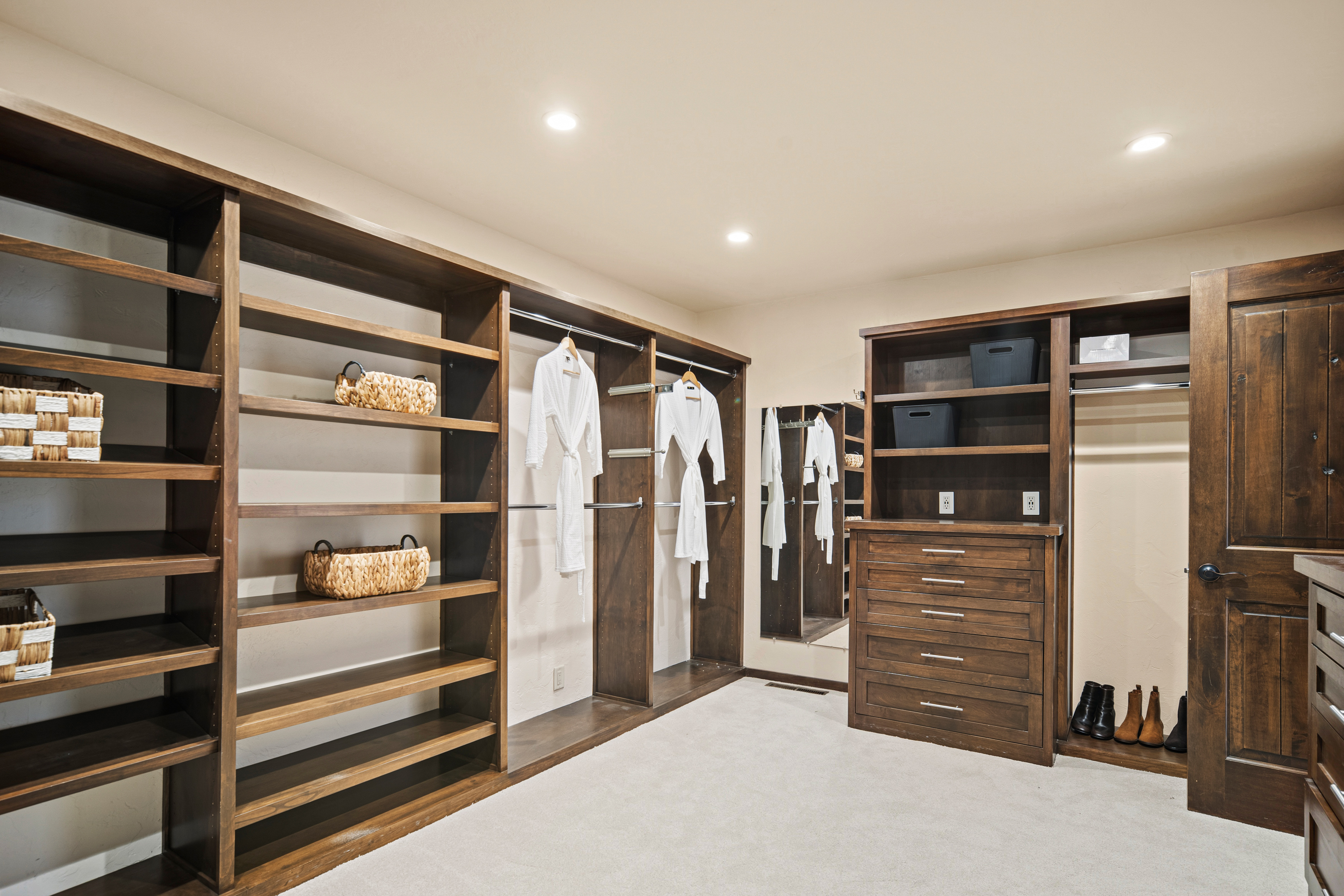 This spacious walk-in closet features extensive dark wood shelving and hanging rods, providing ample storage and organization. The room includes a built-in dresser with multiple drawers, a mirror, and a neutral carpeted floor, creating a clean and functional aesthetic. The perspective is a wide-angle shot from the entrance, showcasing the organized layout and warm, inviting lighting.
