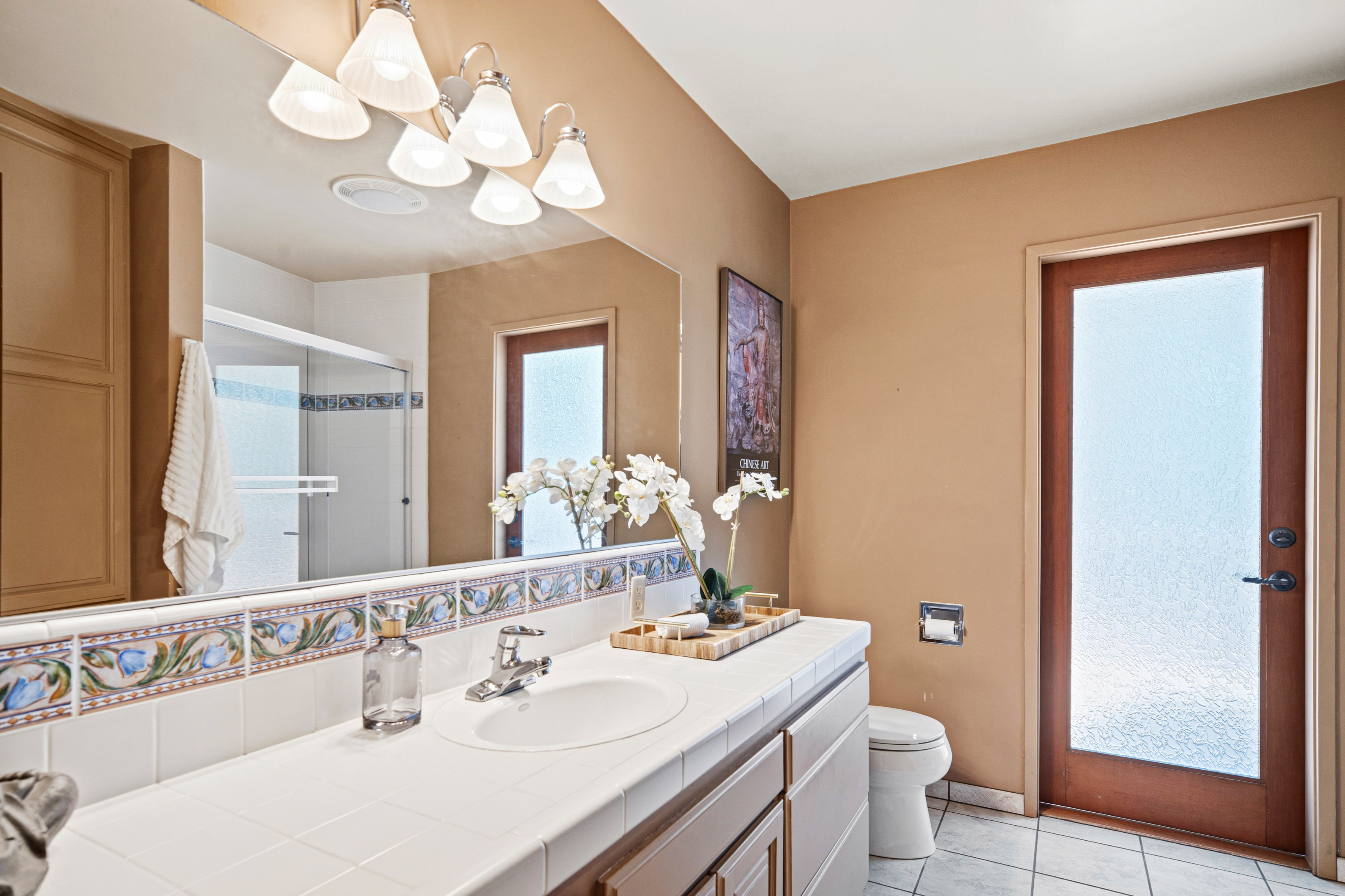 This bathroom features a long vanity with a white tiled countertop and a single sink, accented by a decorative tile backsplash. A large, wall-to-wall mirror reflects the space, including the shower area and a door with frosted glass that leads outside. The room is painted in a warm, neutral tone and includes a toilet and a small decorative tray with artificial flowers on the vanity.