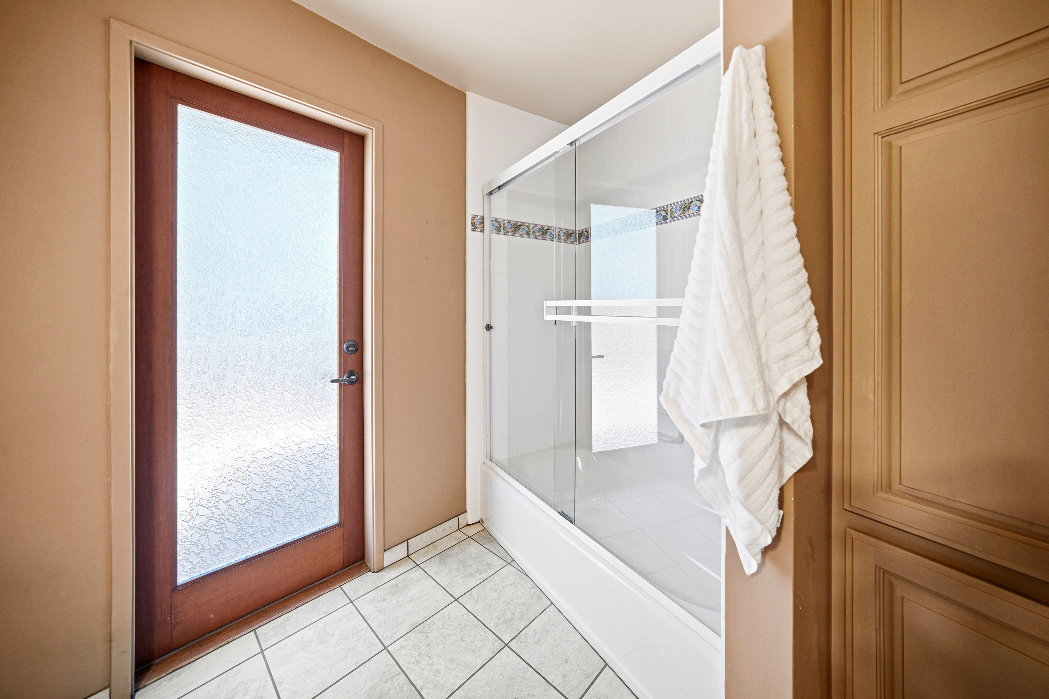 This interior view captures a functional bathroom space featuring a glass-enclosed shower with a sliding door and a decorative tile border. A white towel hangs prominently on the right, while a door with frosted glass leads to an adjacent area, providing a sense of depth. The room is finished with light-colored floor tiles and neutral-toned walls, creating a clean and straightforward aesthetic.
