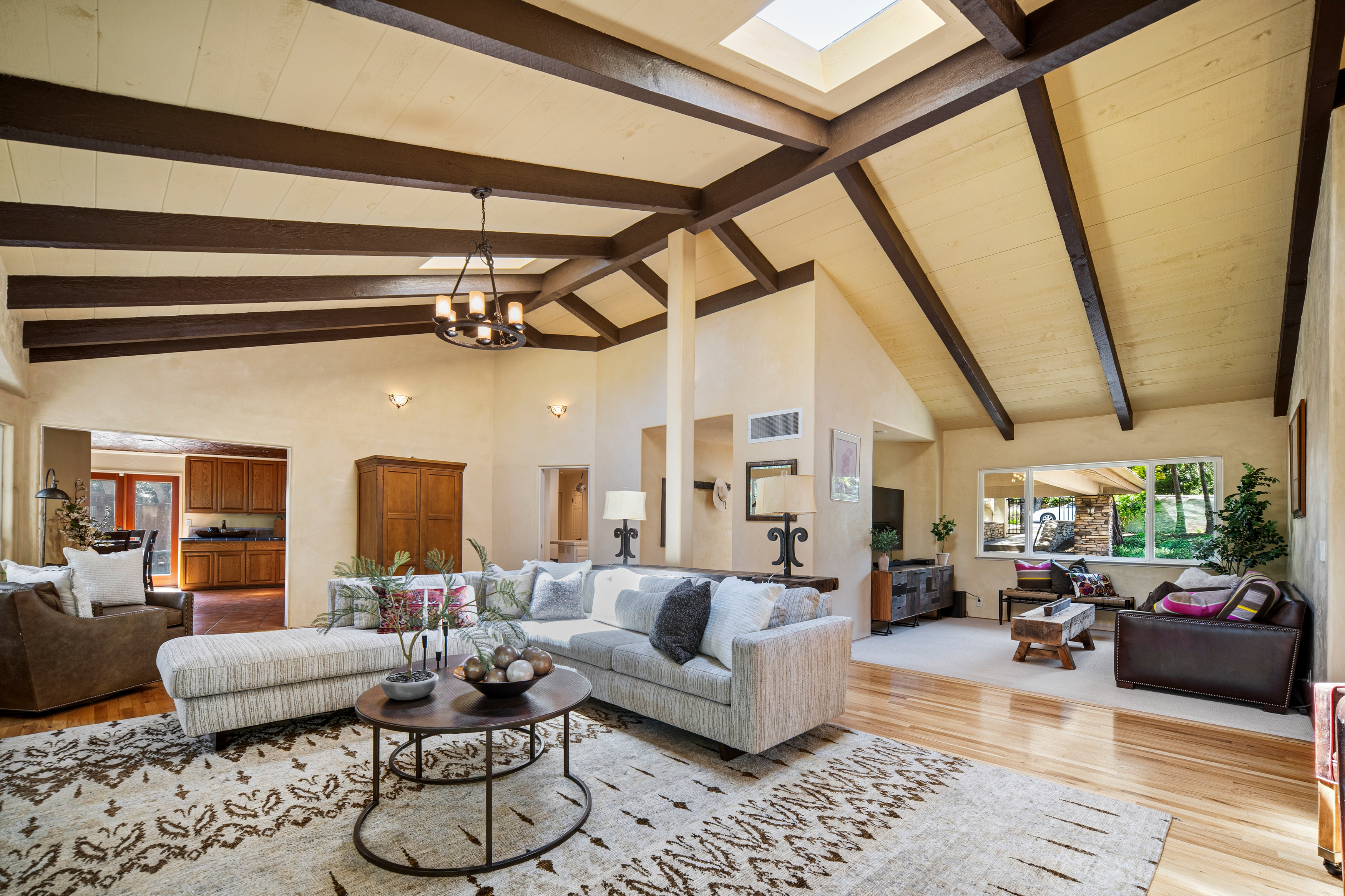 This spacious, open-concept living area features vaulted ceilings with exposed dark wood beams and a central skylight that floods the room with natural light. The space is anchored by a large, light-toned sectional sofa on a patterned area rug, with a secondary seating area visible in the background near a large window. The warm, neutral color palette and hardwood flooring create an inviting, airy atmosphere that flows seamlessly into the adjacent kitchen and secondary living space.