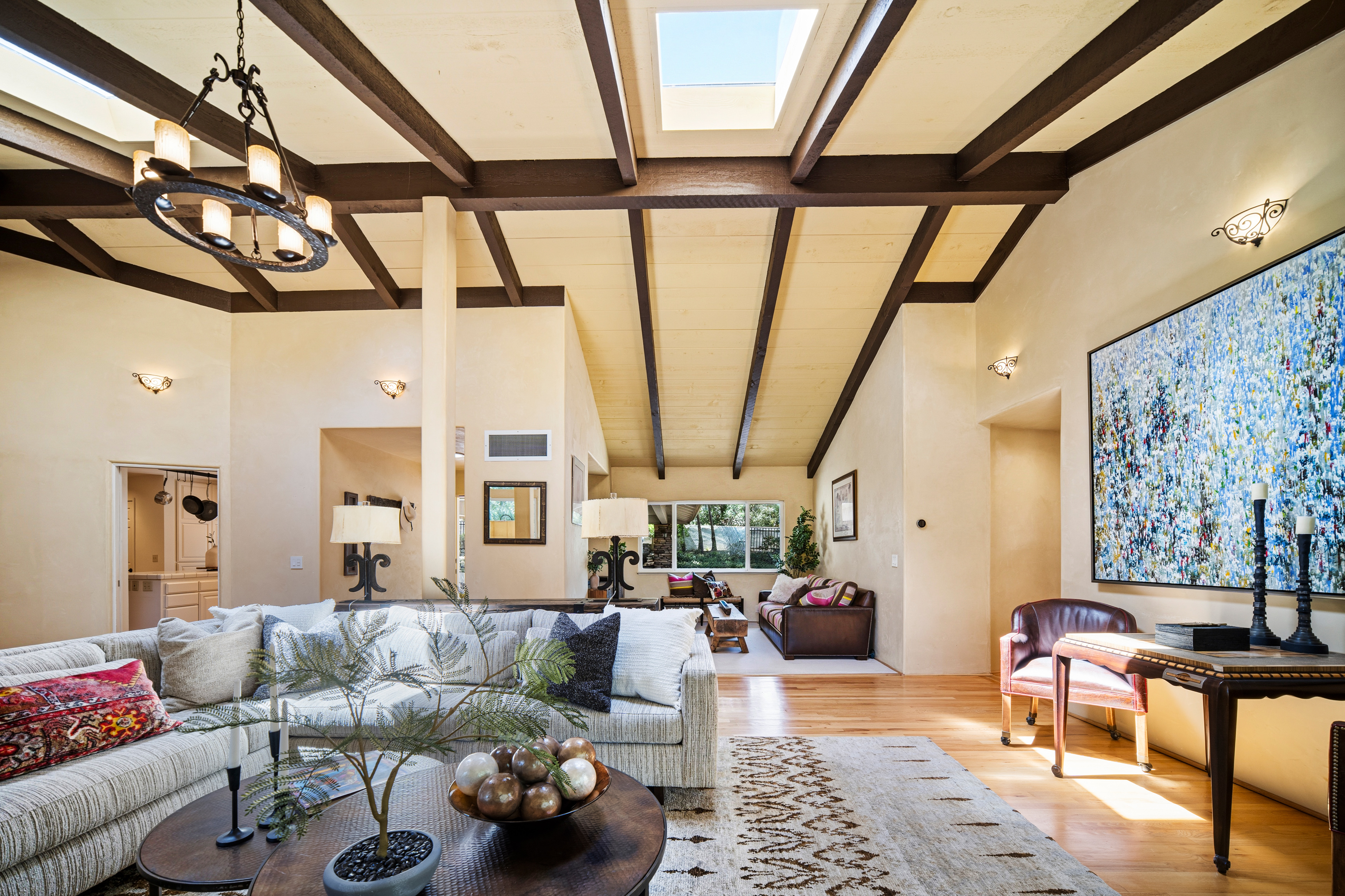 This spacious, open-concept living area features vaulted ceilings with exposed dark wood beams and multiple skylights that flood the room with natural light. The space is anchored by a large, neutral-toned sectional sofa, a round coffee table with a decorative bowl, and a patterned area rug over light wood flooring. A large, vibrant abstract painting serves as a focal point on the wall, while a secondary seating area is visible in the background, creating a warm and inviting atmosphere.
