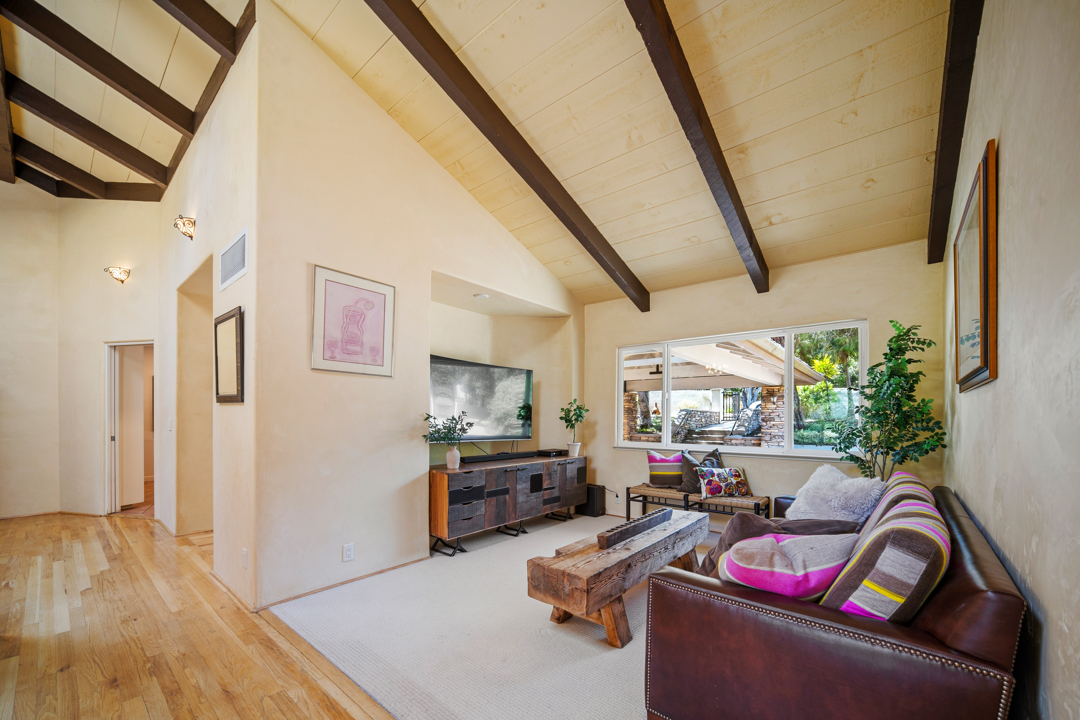 This inviting living room features a vaulted ceiling with exposed dark wood beams and warm, neutral-toned walls. The space is anchored by a leather sofa with colorful accent pillows and a rustic, heavy wood coffee table, while a unique, multi-textured media console sits beneath a wall-mounted television. Large windows provide a bright view of the outdoor patio area, creating a seamless connection between the indoor living space and the exterior landscape.