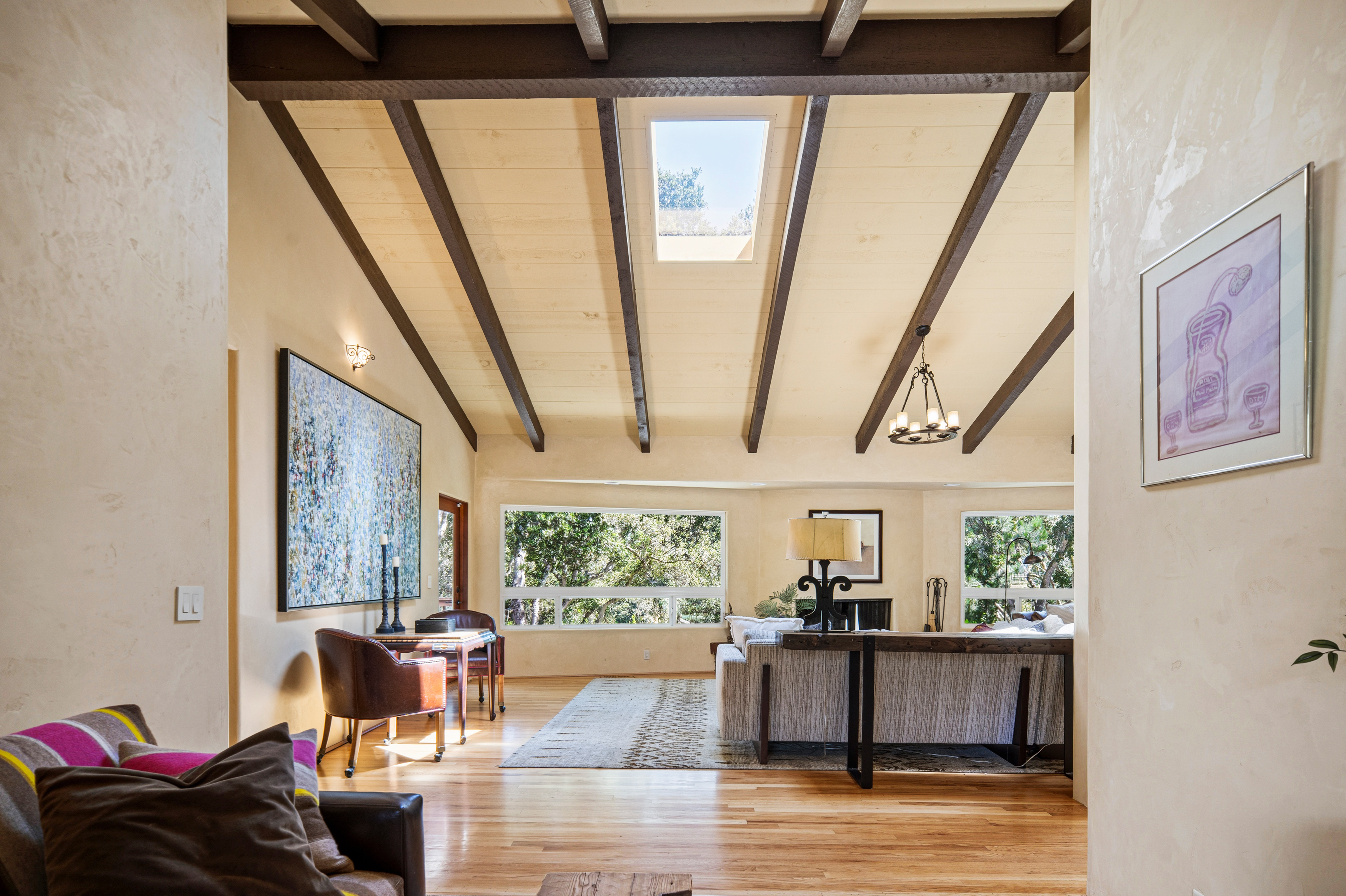 This spacious living area features a vaulted ceiling with exposed dark wood beams and a central skylight that floods the room with natural light. The space is anchored by a large, textured area rug and a comfortable sofa, complemented by a small desk area and a large abstract painting on the wall. Large windows provide a scenic view of the surrounding trees, creating a bright and airy atmosphere with a warm, inviting aesthetic.