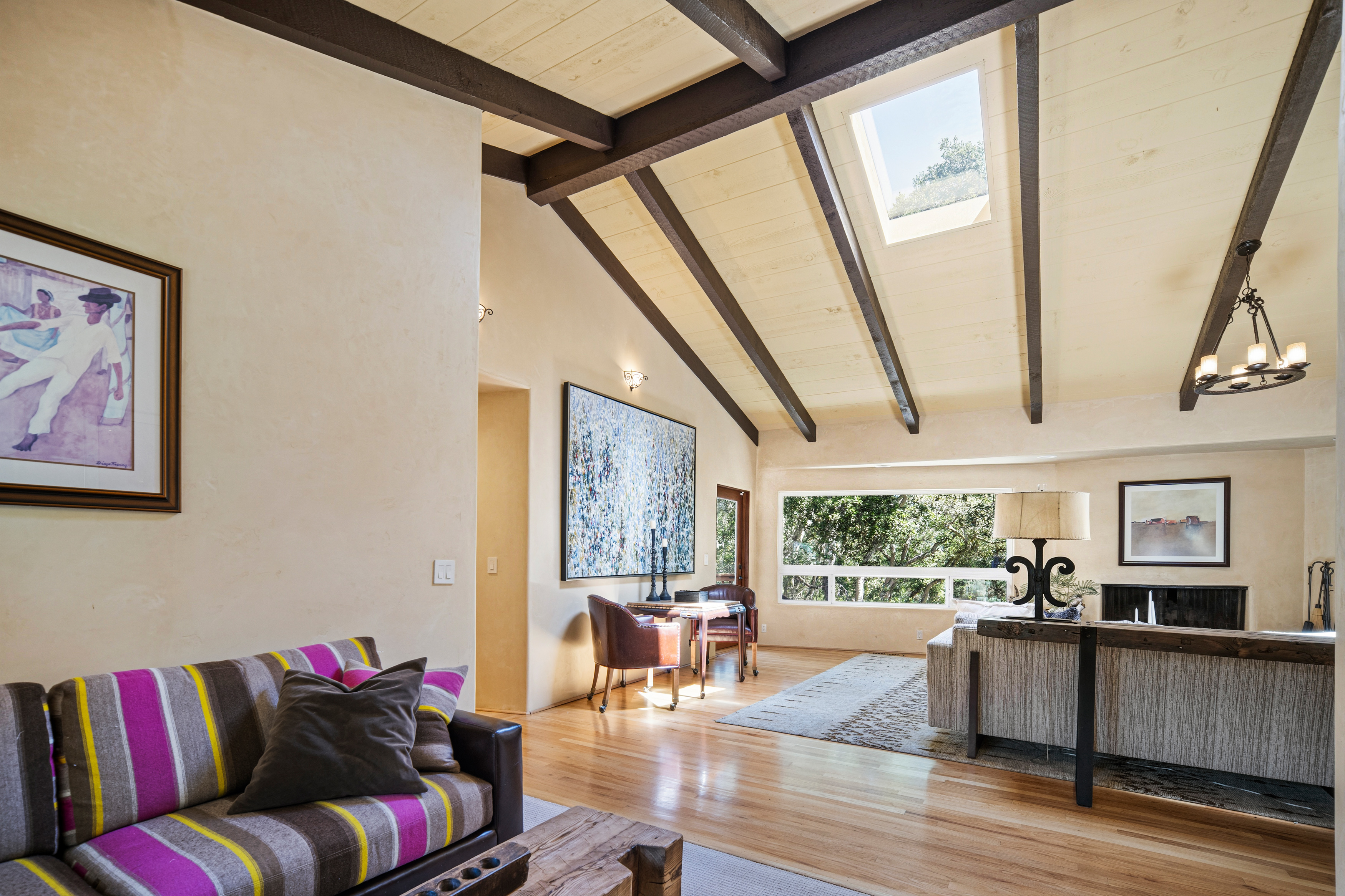 This spacious living room features a vaulted ceiling with exposed dark wood beams and a skylight that fills the space with natural light. The room is anchored by a striped sofa in the foreground and a cozy seating area near a fireplace, with large windows offering a view of the surrounding trees. The warm wood flooring and neutral walls create an inviting, open-concept atmosphere perfect for relaxation.