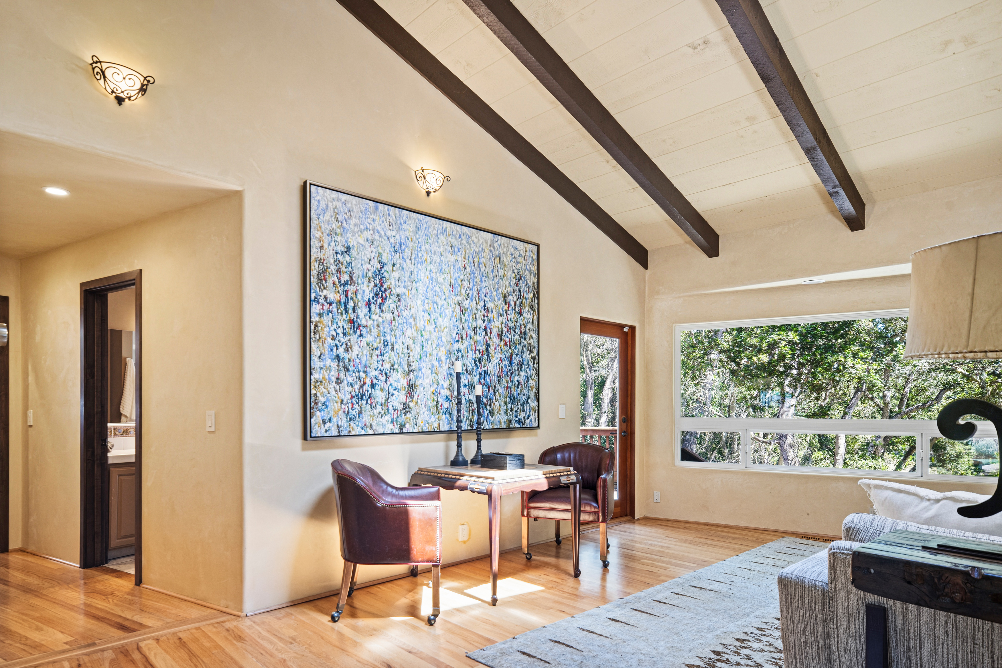 This bright living area features vaulted ceilings with exposed dark wood beams and warm, neutral-toned walls. A large, vibrant abstract painting serves as a focal point above a small wooden table flanked by two leather chairs, while a large window provides a scenic view of the surrounding trees. The space is finished with light wood flooring and a textured area rug, creating a cozy and inviting atmosphere.