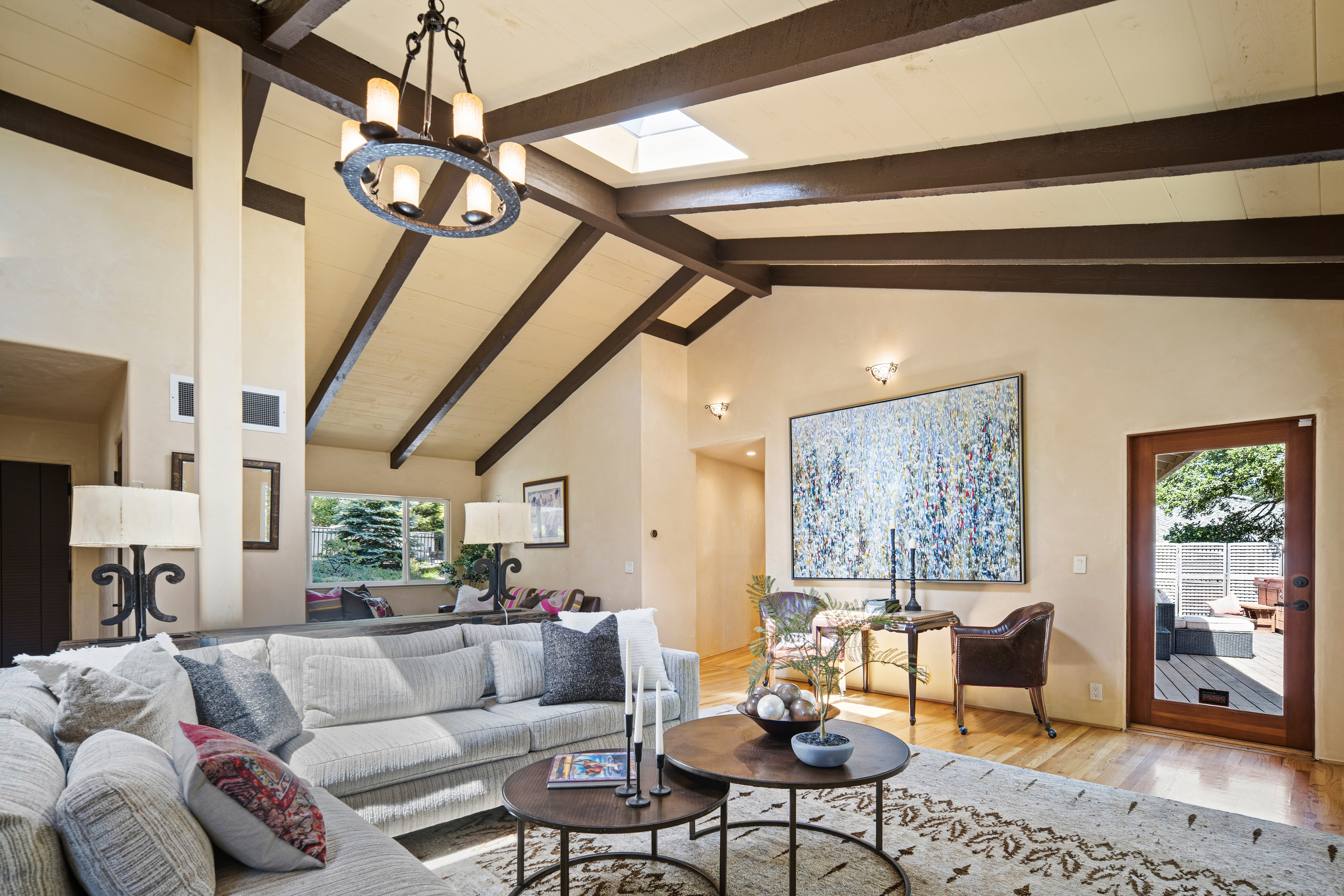 This spacious living room features a dramatic vaulted ceiling with exposed dark wood beams and a central skylight that floods the area with natural light. A large, textured abstract painting serves as a focal point on the wall, complemented by a comfortable light-toned sectional sofa and a pair of nested round coffee tables. The room flows seamlessly toward a glass-paned door that opens onto an outdoor deck, creating an inviting and airy atmosphere.