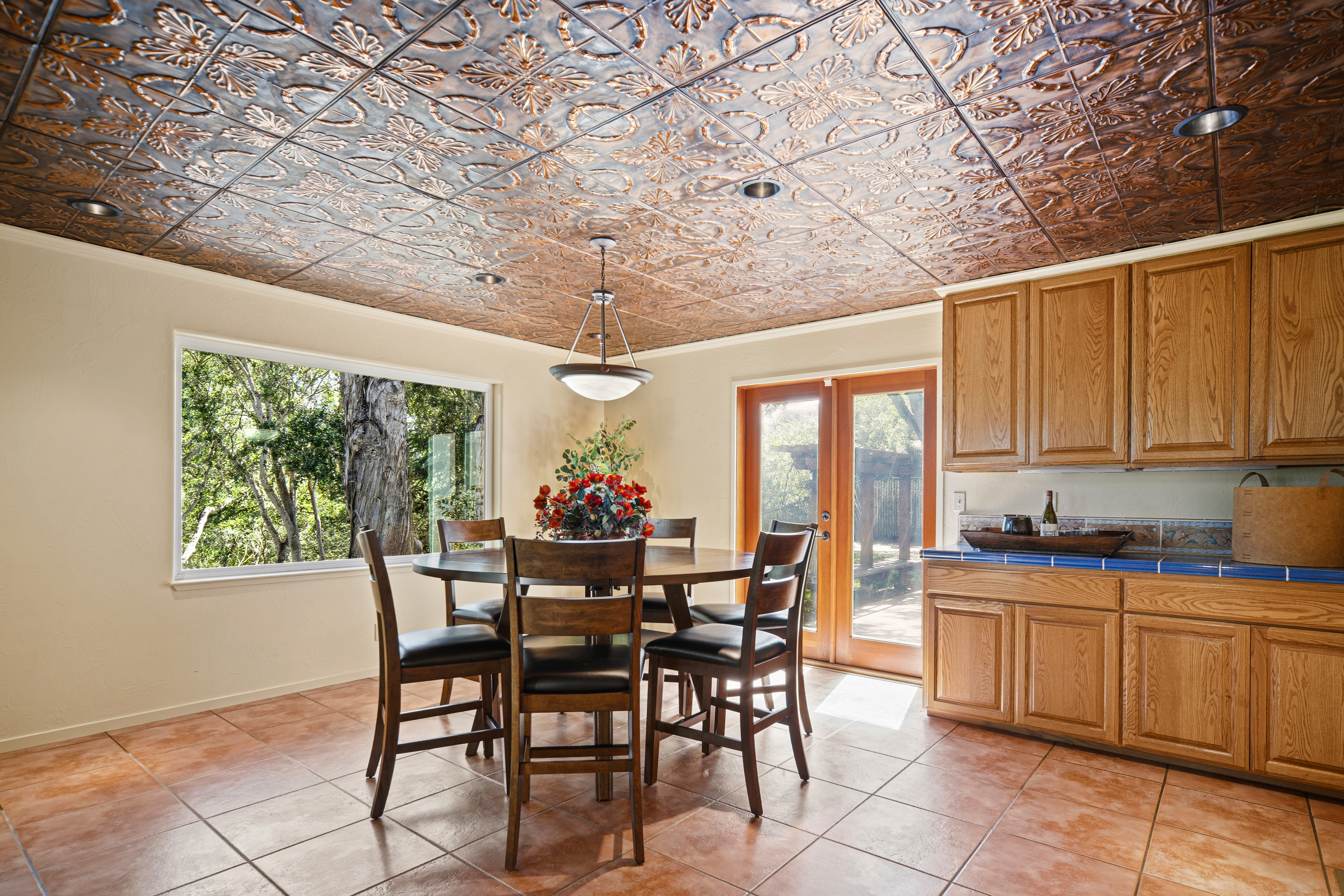 This dining area features a round wooden table surrounded by four chairs, positioned beneath a decorative pendant light. The room is distinguished by a unique, metallic-finish patterned ceiling and warm-toned tile flooring. A large window and a set of French doors provide ample natural light and views of the surrounding greenery, while a built-in wooden cabinet unit offers convenient storage and counter space.
