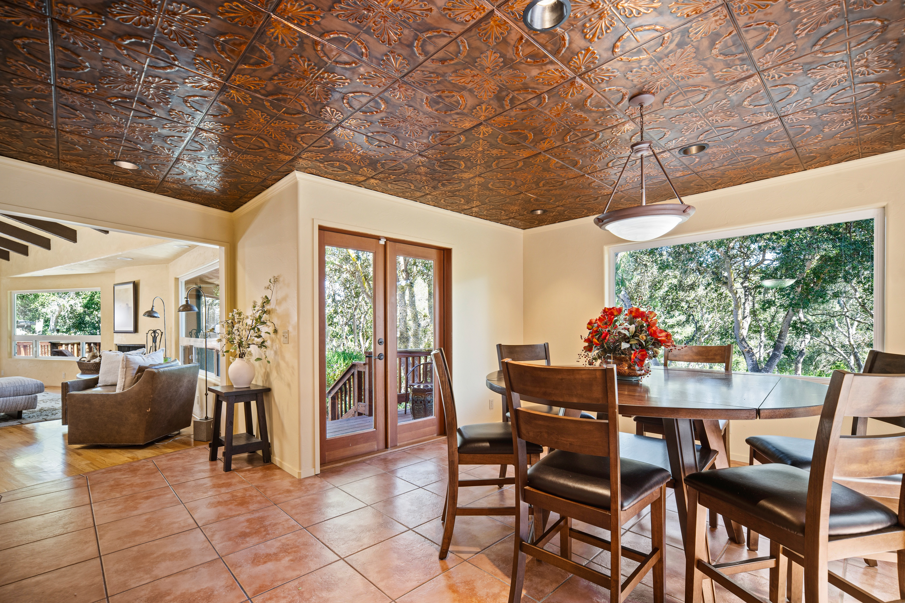 This dining area features a distinctive copper-toned patterned ceiling and warm-toned tile flooring, creating a cozy and rustic atmosphere. A round wooden table with matching chairs sits beneath a modern pendant light, while large windows provide a scenic view of the surrounding trees. The space flows seamlessly into an adjacent living area, highlighting an open and inviting floor plan.