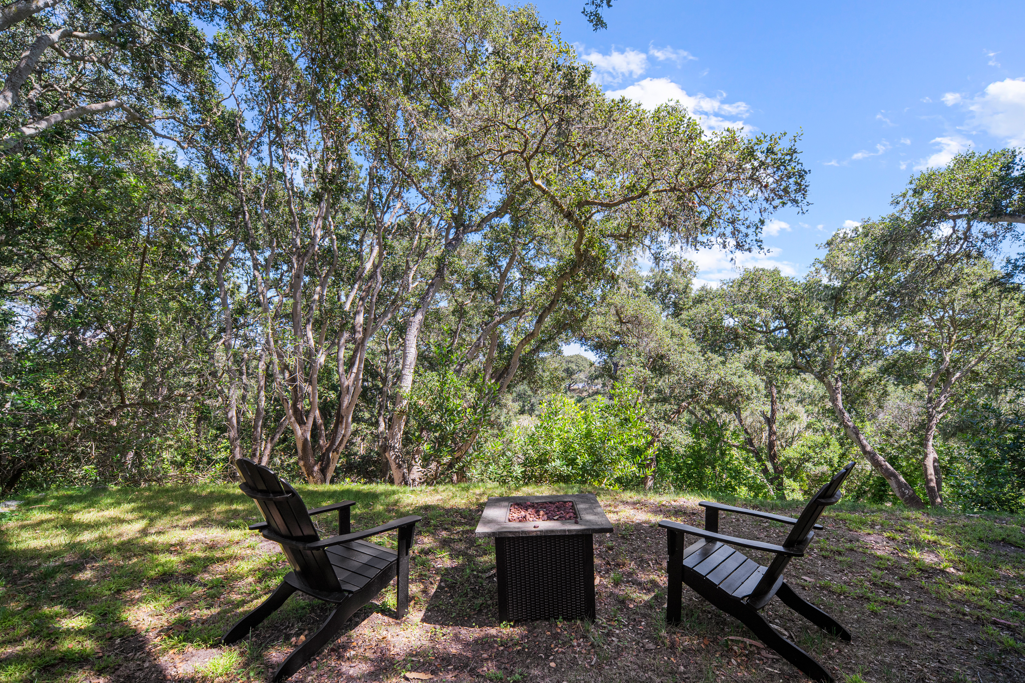 This serene outdoor seating area features two dark Adirondack chairs positioned around a square, dark-wicker fire pit filled with lava rocks. The setting is nestled within a lush, wooded landscape, providing a private and tranquil atmosphere. The perspective is a straight-on, eye-level shot that captures the inviting nature of this secluded backyard retreat.