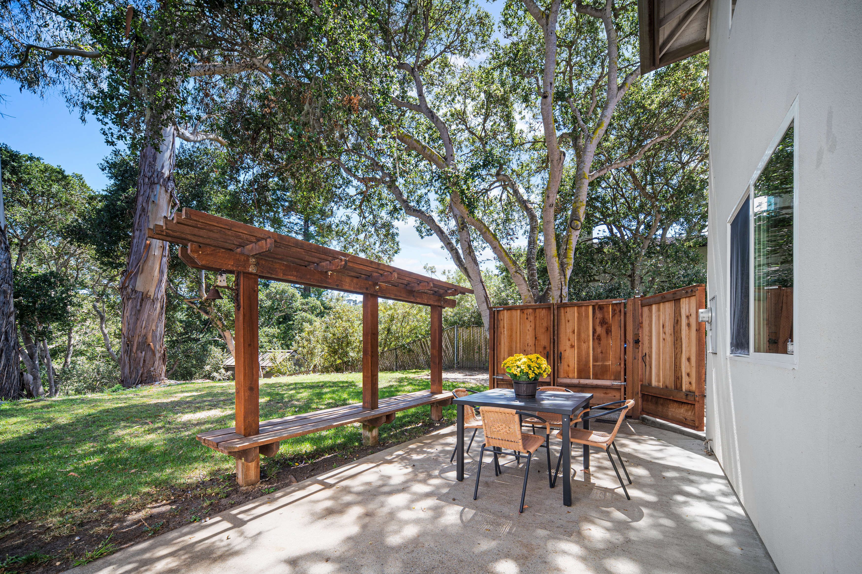 This outdoor patio space features a concrete slab dining area furnished with a table and four wicker-style chairs, accented by a potted yellow floral arrangement. Adjacent to the dining area is a wooden pergola structure with an integrated bench, providing a shaded spot overlooking a grassy yard and mature trees. The scene is framed by a wooden privacy fence and the side of the home, creating a peaceful and private atmosphere for outdoor relaxation.