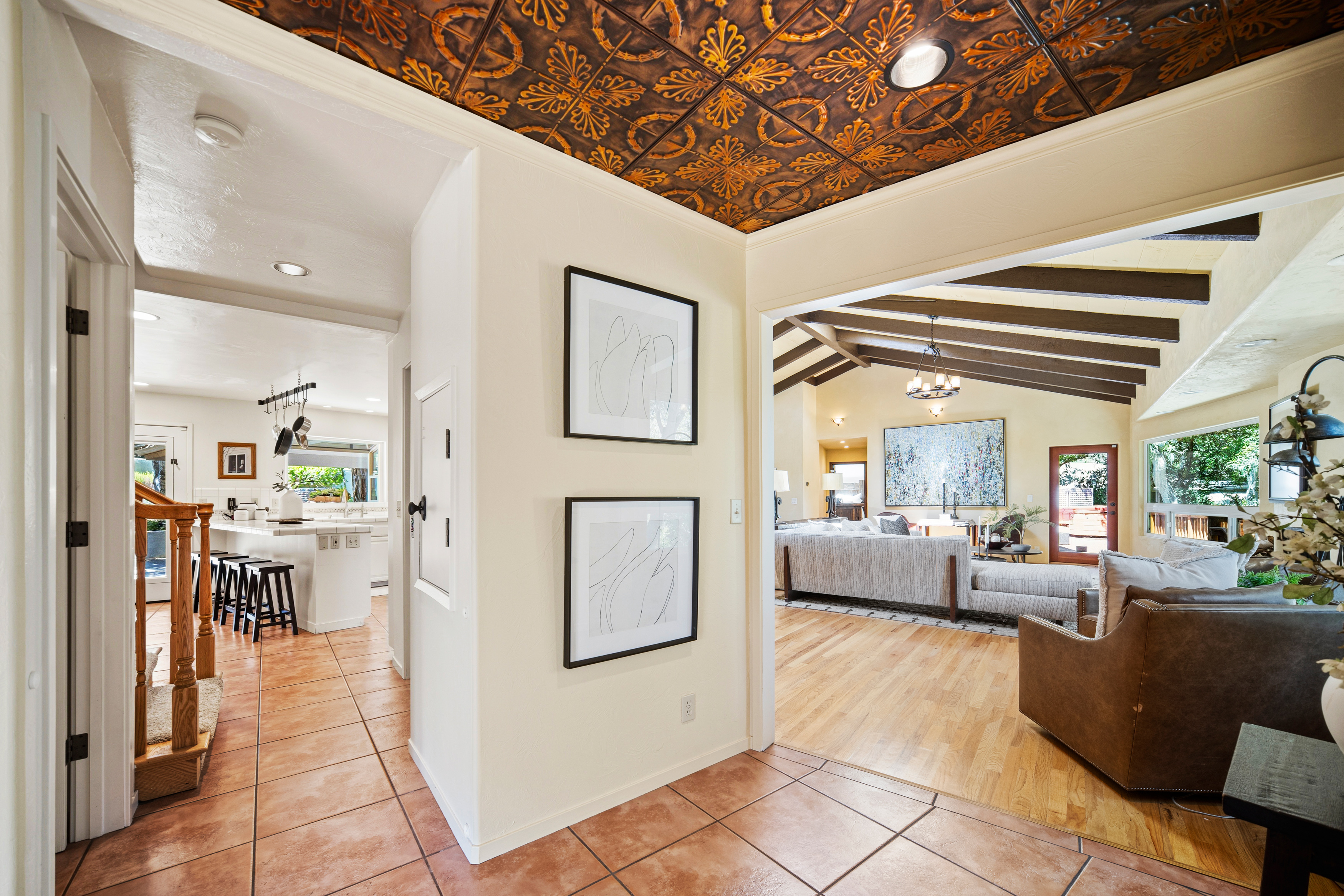 This perspective captures a central foyer area that serves as a transition point between the kitchen, living room, and staircase. The space features a striking decorative metal ceiling with a floral pattern, contrasting with the light-toned walls and warm-toned tile flooring. Two framed abstract art pieces hang on the central wall, while the open layout provides a clear view into the bright, wood-floored living area with exposed ceiling beams.