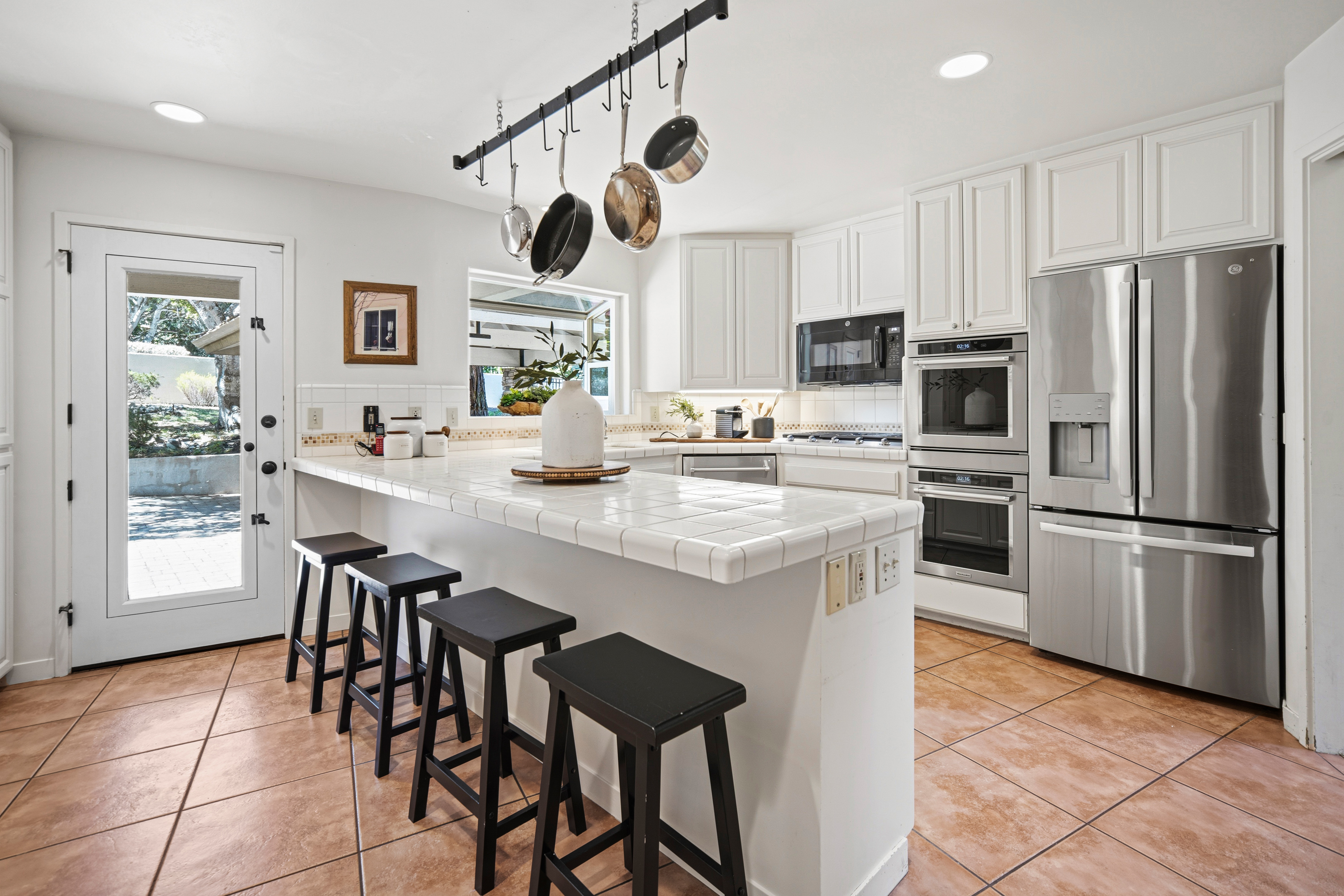 This bright kitchen features white cabinetry, a tiled countertop island with four black bar stools, and stainless steel appliances including a double wall oven and a refrigerator. A hanging pot rack above the island adds a functional, rustic touch, while the terracotta-toned floor tiles provide a warm contrast to the neutral walls. The space is well-lit by recessed ceiling lights and a window that looks out toward an outdoor patio area.