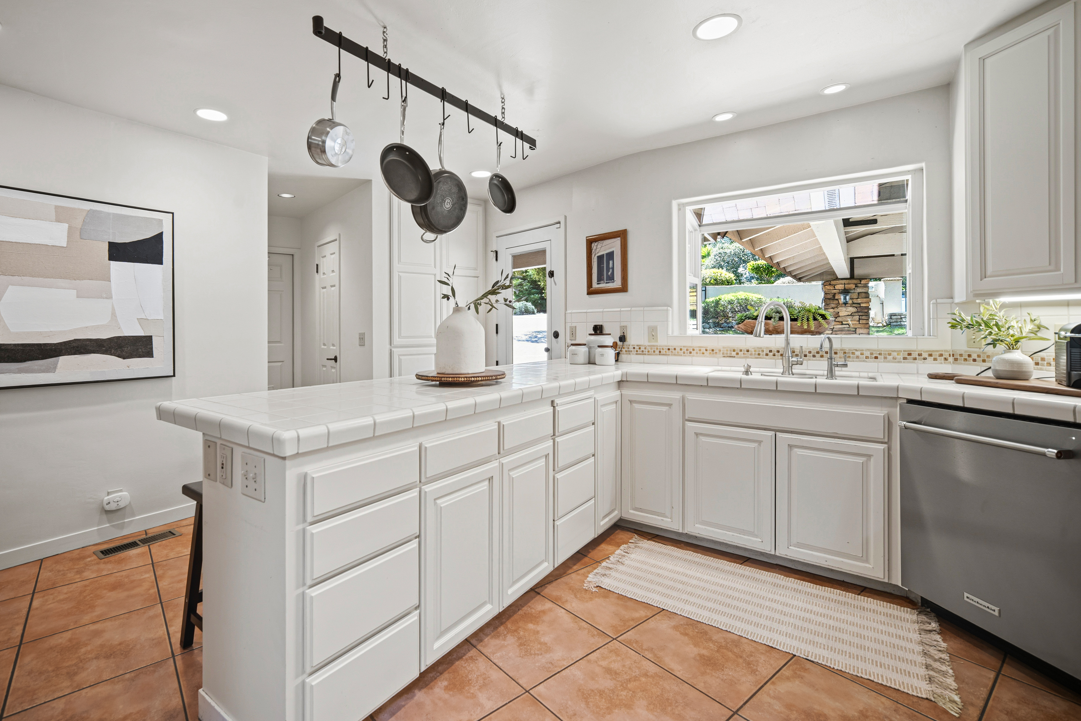 This bright kitchen features white cabinetry paired with tiled countertops and a matching tiled backsplash. A central island provides additional workspace and seating, while a hanging rack above holds cookware, adding a functional touch to the space. The room is finished with warm-toned floor tiles and a large window above the sink that looks out toward the exterior landscape.