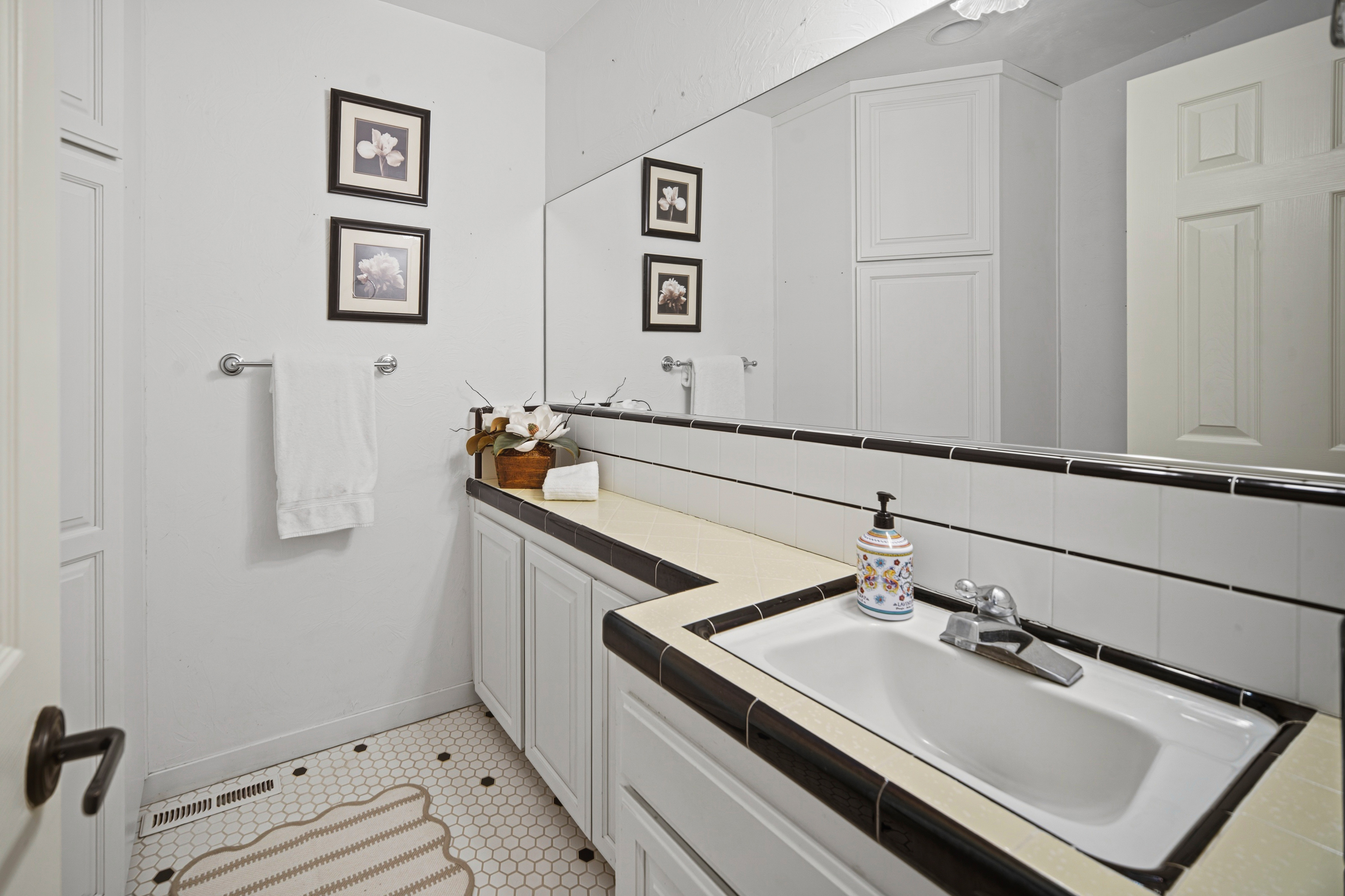 This bathroom features a classic aesthetic with a white vanity topped with light-colored tile and a dark tile border. A large wall-to-wall mirror reflects the space, while the floor is finished in a white hexagonal tile with dark accent dots. The room is decorated with framed floral artwork and a white towel hanging on a wall-mounted rack.